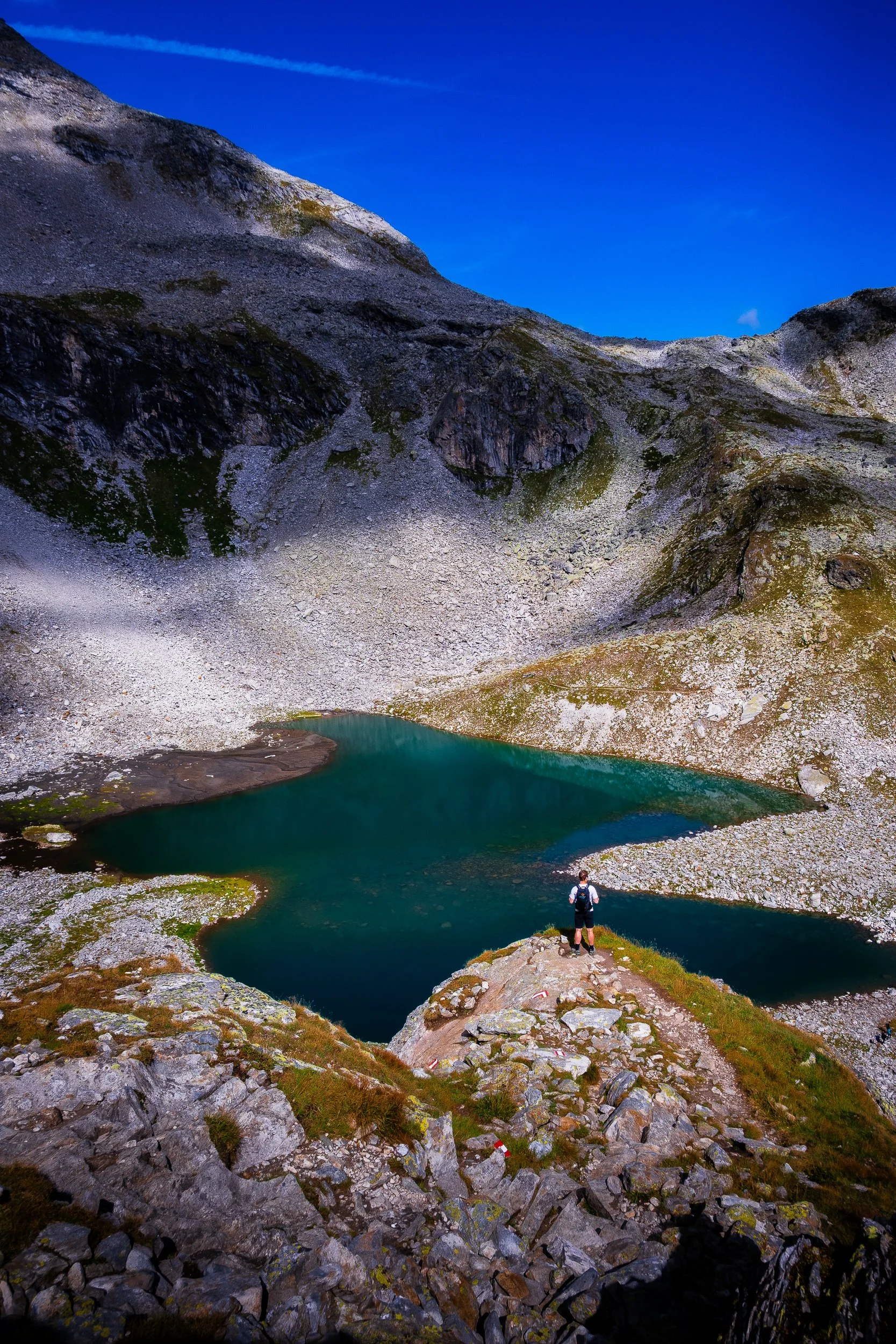 Hiker overlooking a turquoise alpine lake surrounded by rocky mountain slopes under a deep blue sky