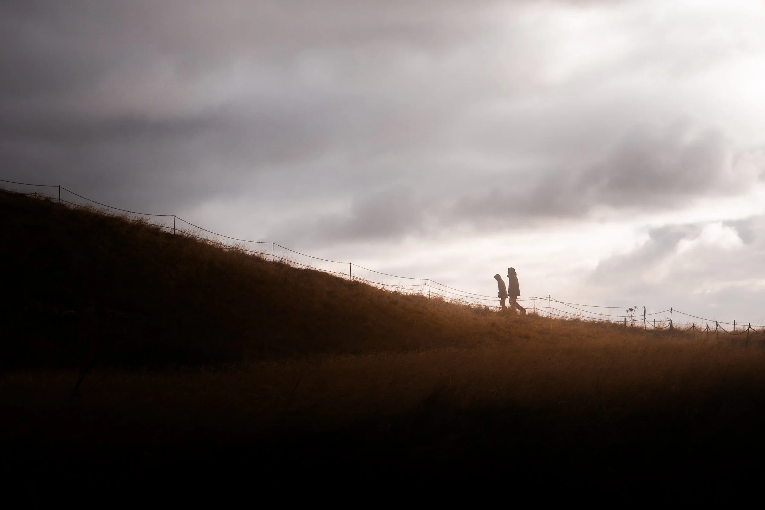 Two people walking along a grassy hillside beneath a dramatic cloudy sky