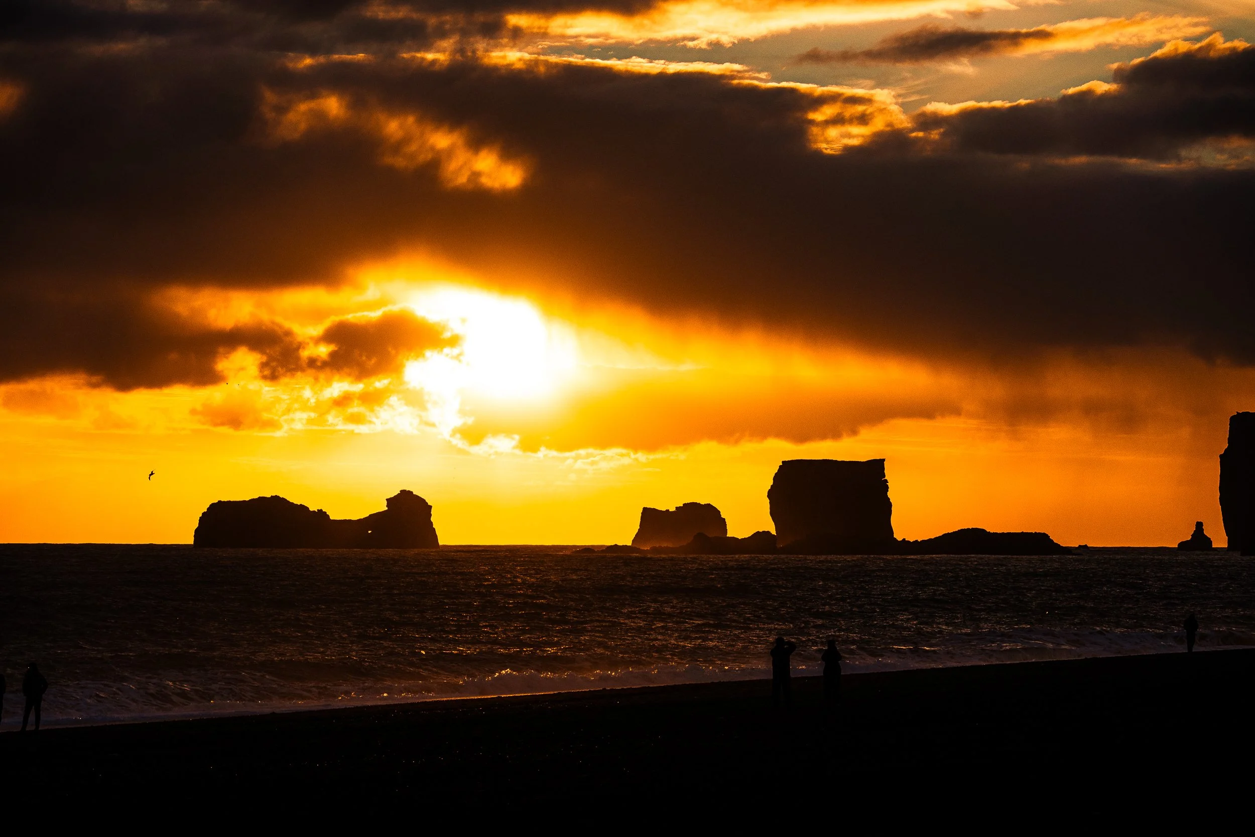 Dramatic golden sunset over ocean sea stacks, dark clouds, waves, and silhouetted people on a beach