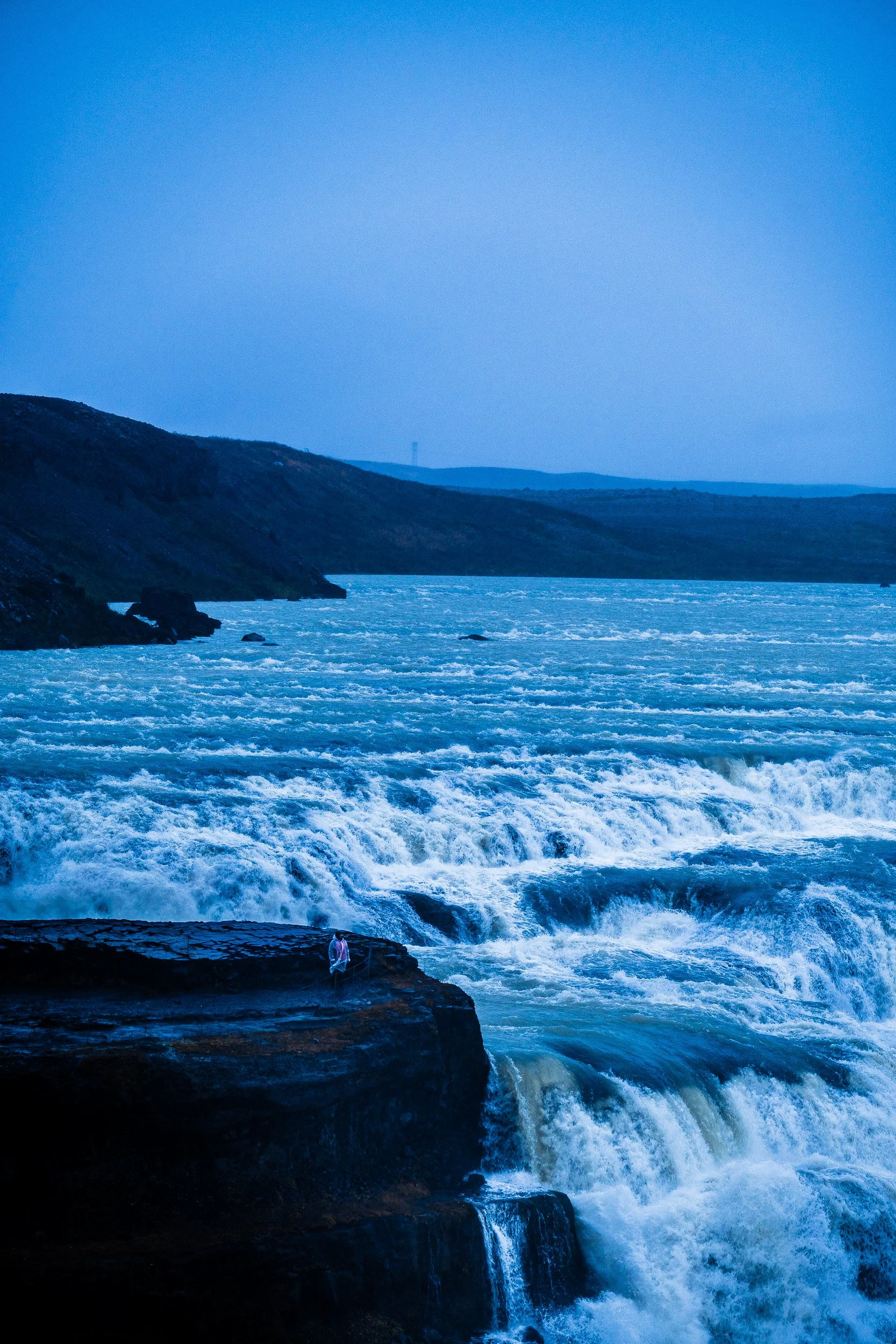 Powerful ocean waves crashing against dark rocky cliffs beneath a blue sky, with a person standing near the edge.