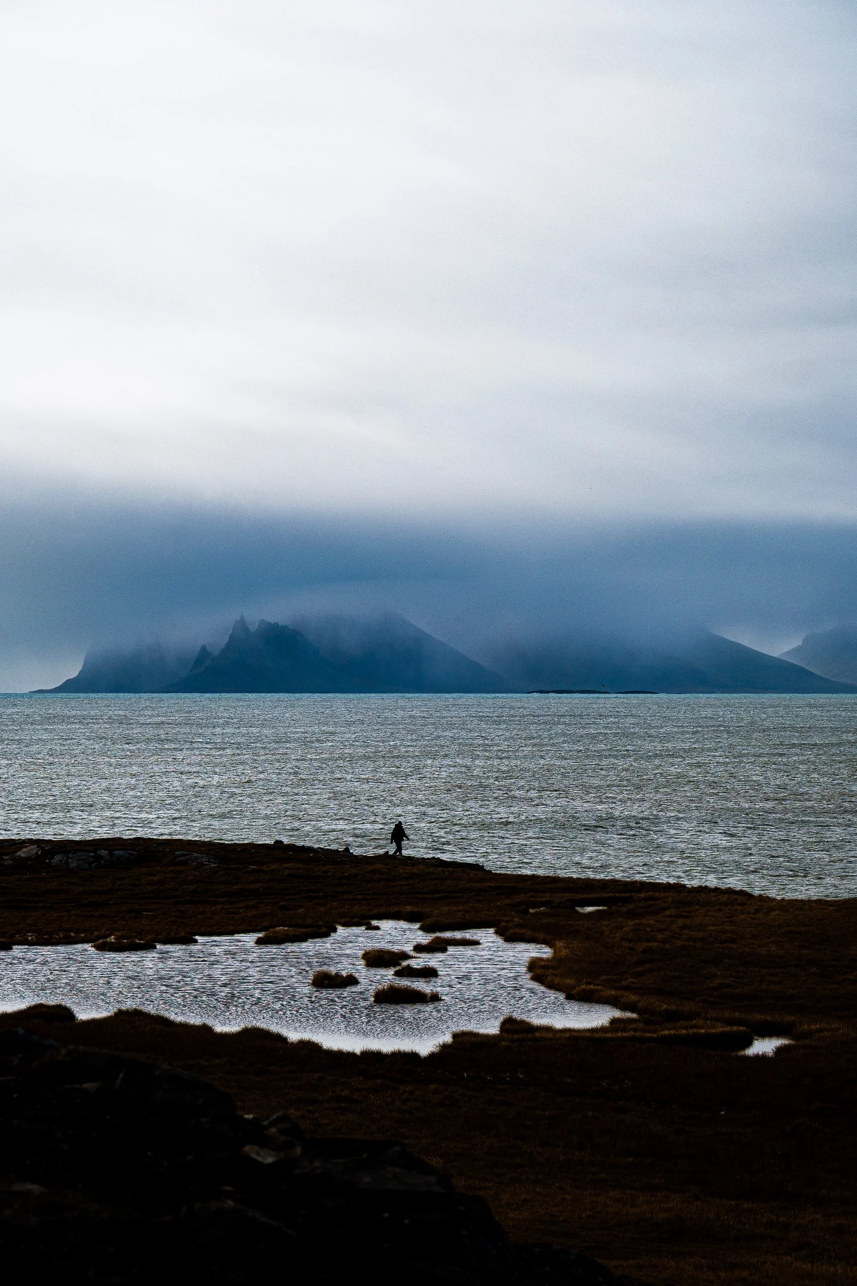 Silhouetted hiker beside a coastal lagoon with rugged mountains shrouded in fog