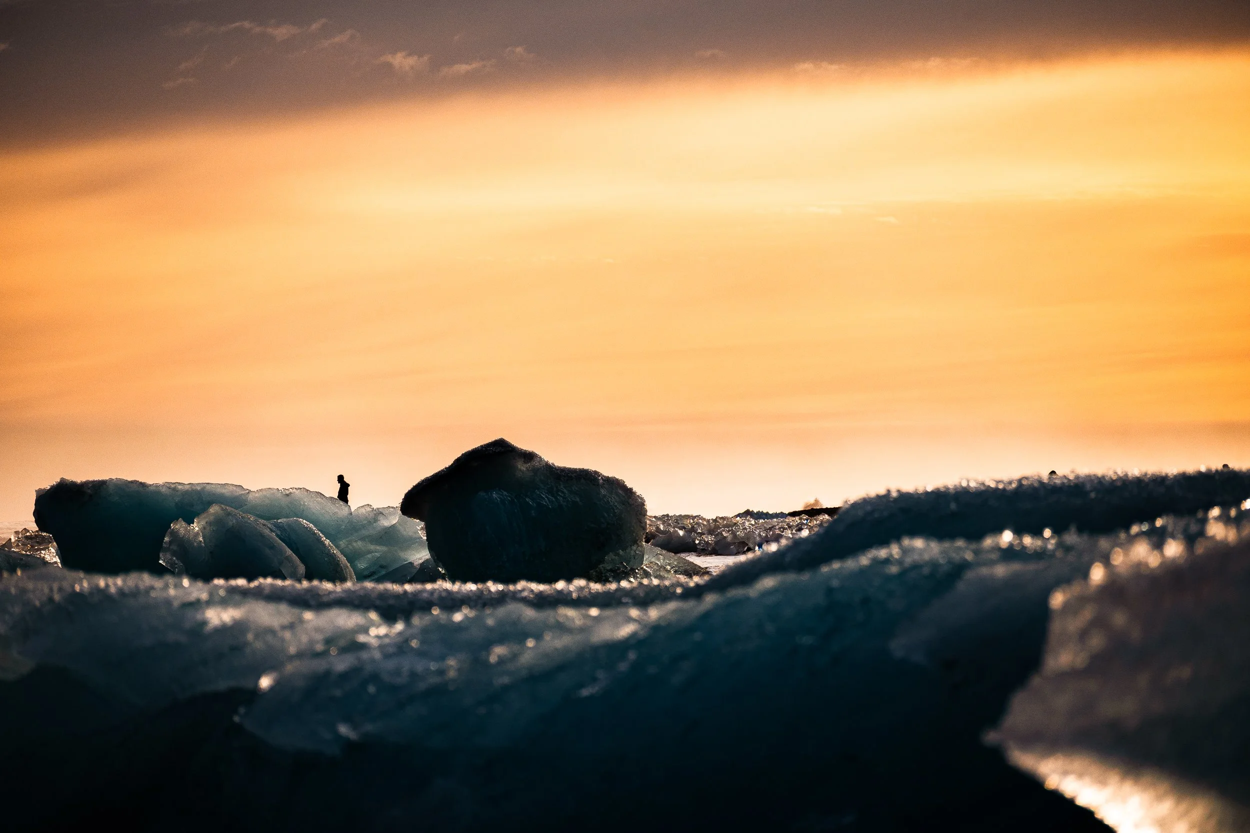 Ice chunks on a dark shoreline silhouetted against a glowing orange sunset sky