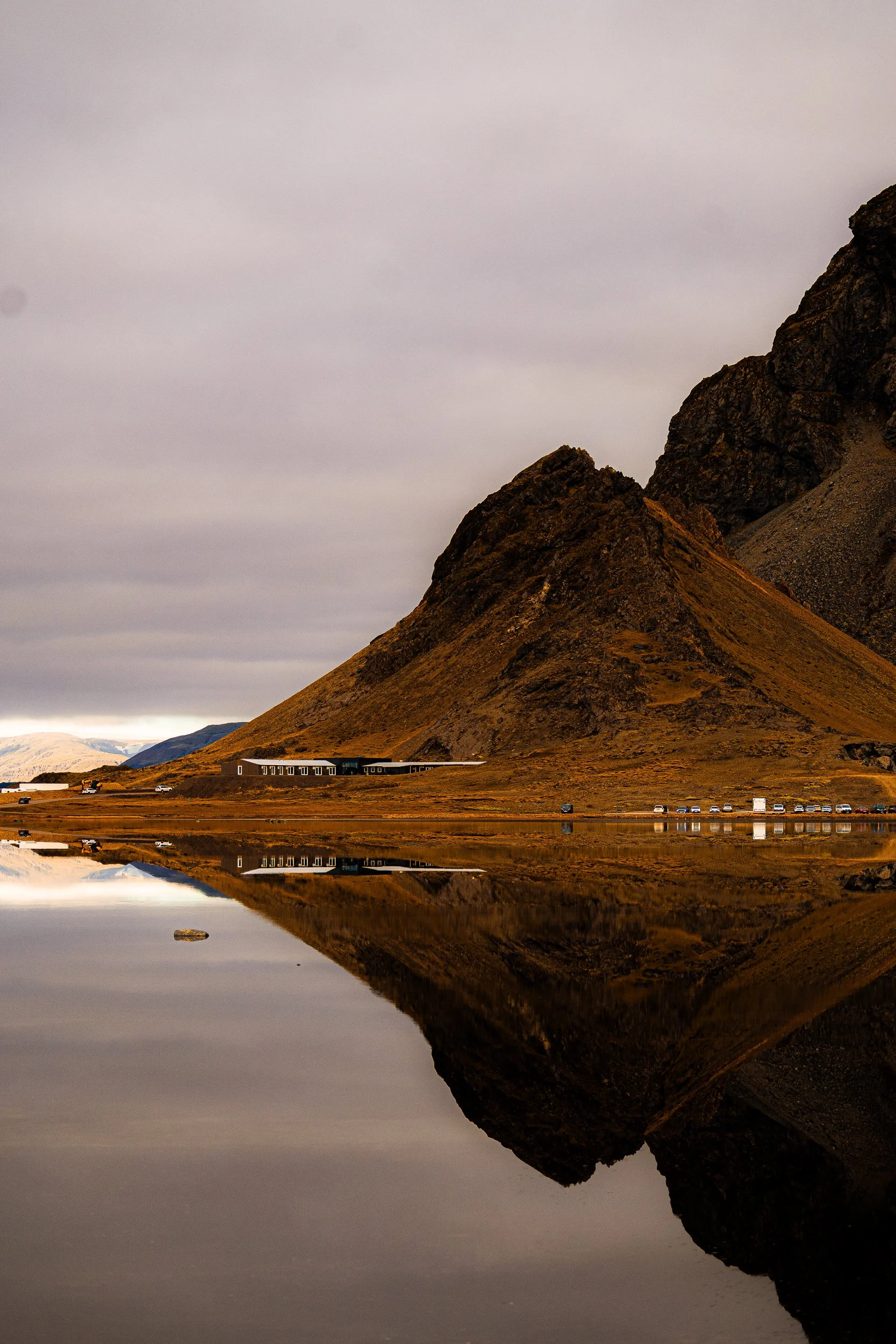 Rugged mountain and lakeside building reflected in a calm pond beneath an overcast sky
