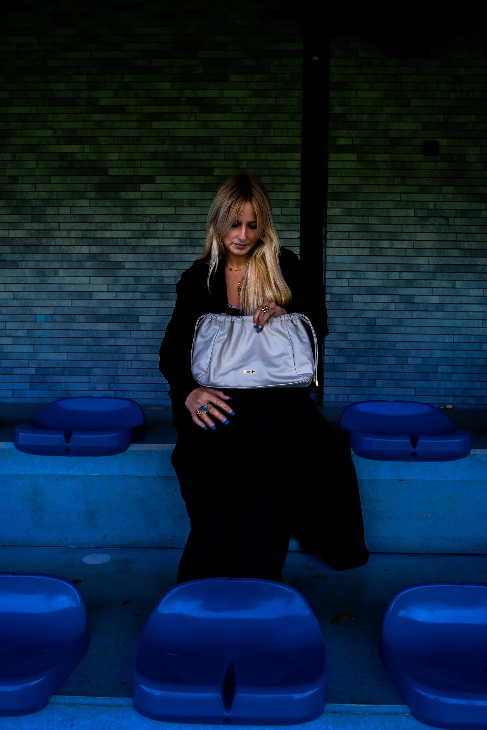 Blonde woman in black clothing holding a white handbag while seated among blue stadium seats.