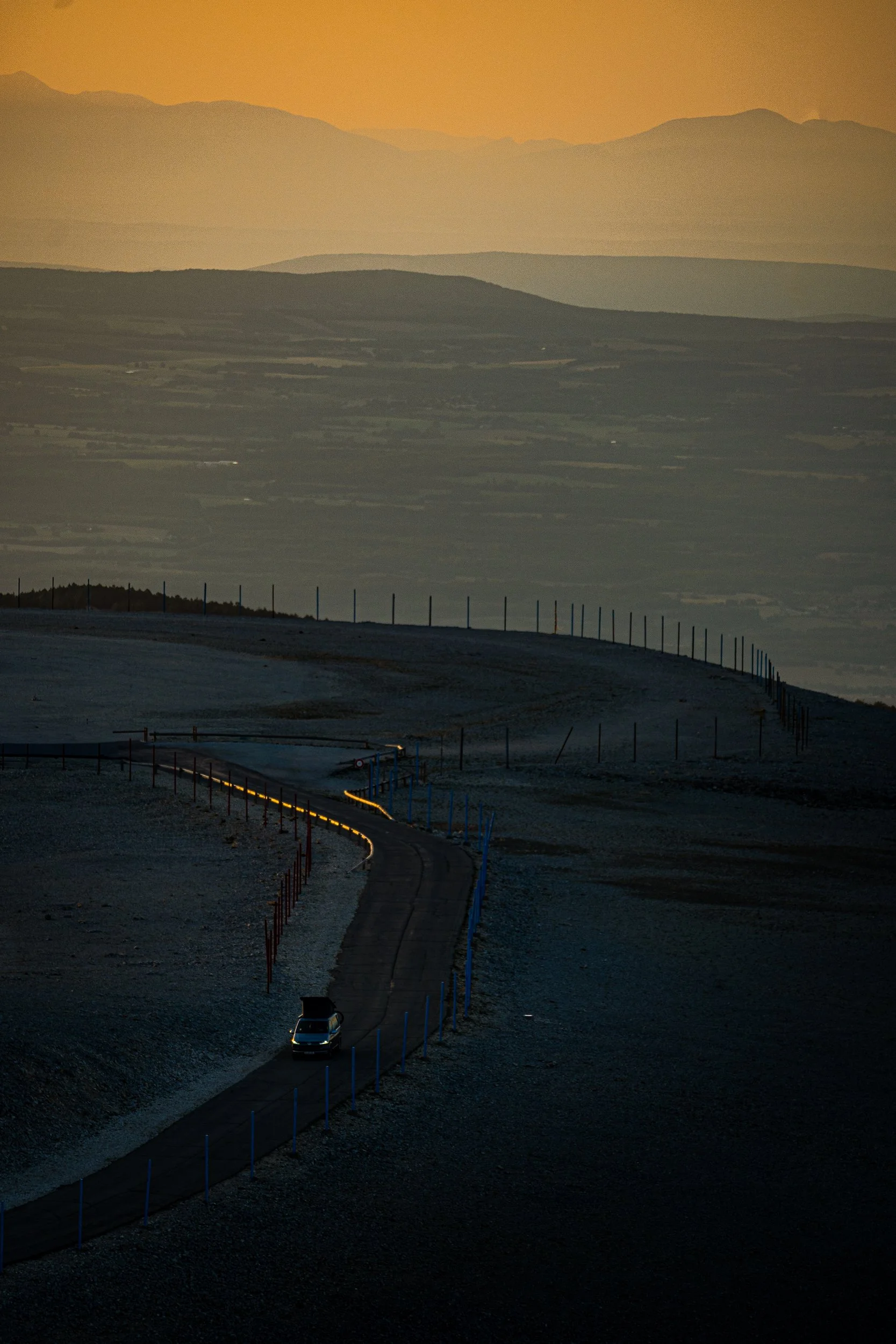 A car travels along a winding mountain road through a frosty landscape at sunset, with layered mountain ranges in the background.