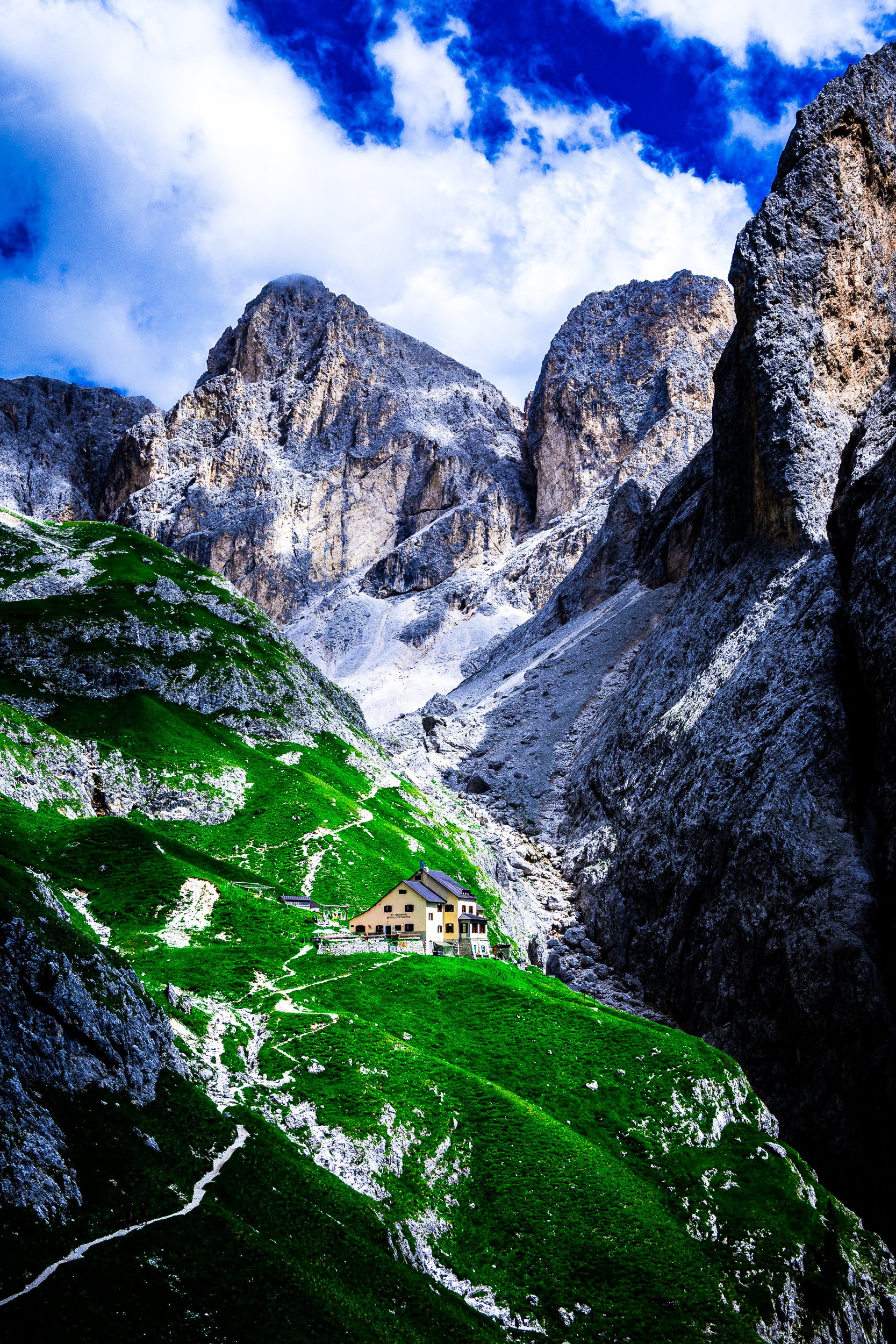 Mountain hut surrounded by vivid green alpine meadows and dramatic rocky peaks beneath a cloudy blue sky