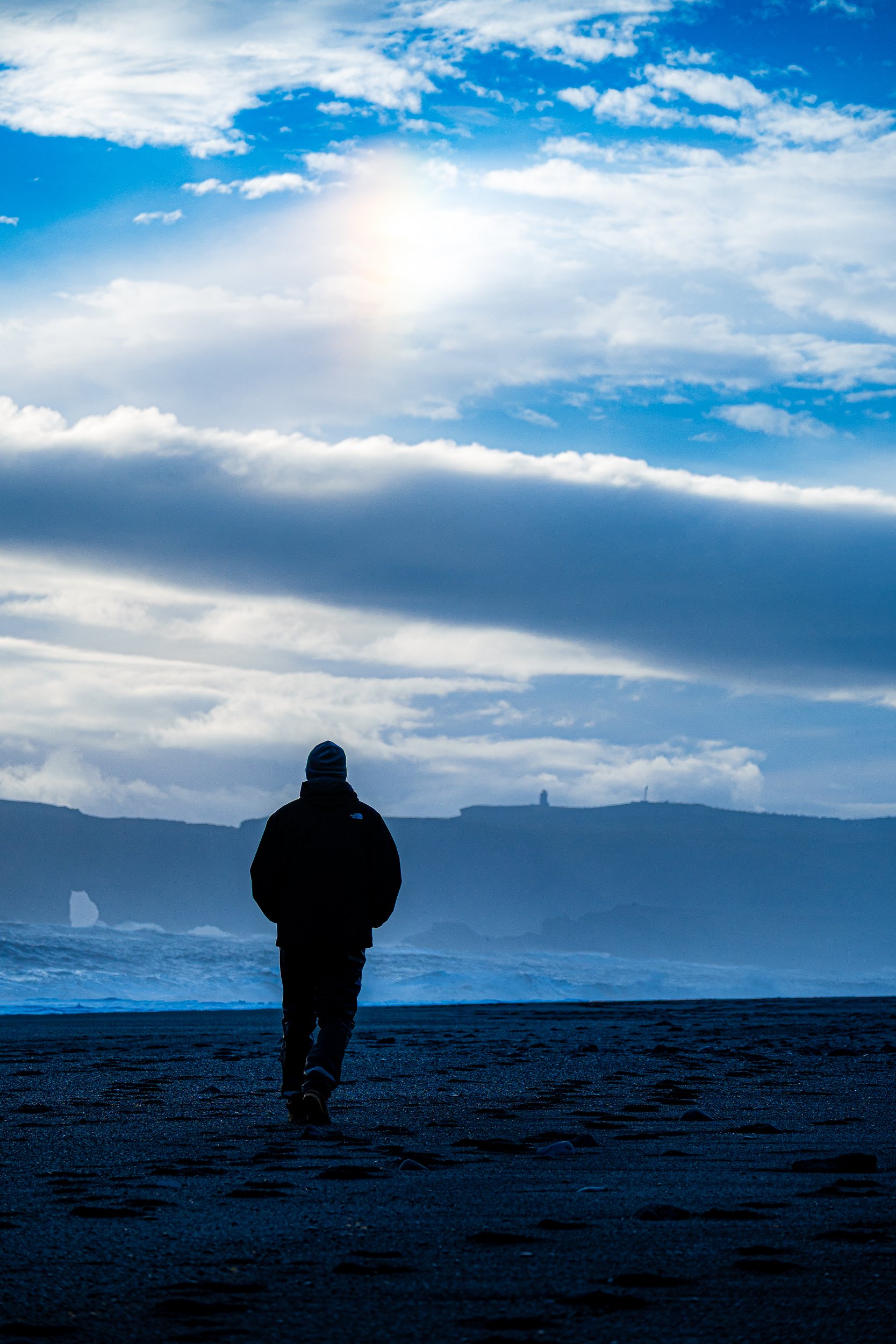 Person in a black jacket walking across a volcanic beach beneath a bright, cloud-filled sky