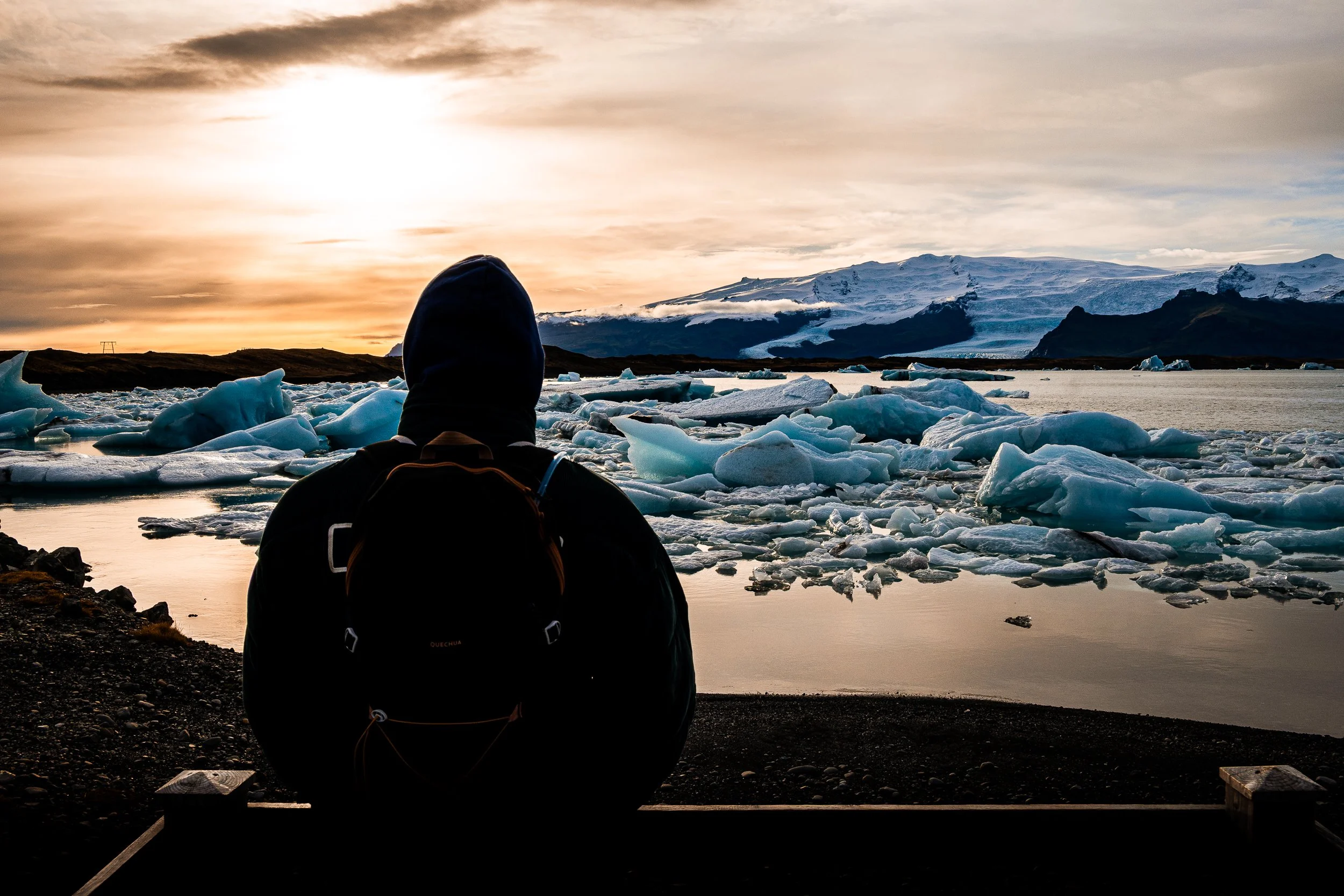Hiker with a backpack viewing floating icebergs in a glacial lagoon at sunset, with a snow-covered mountain behind.