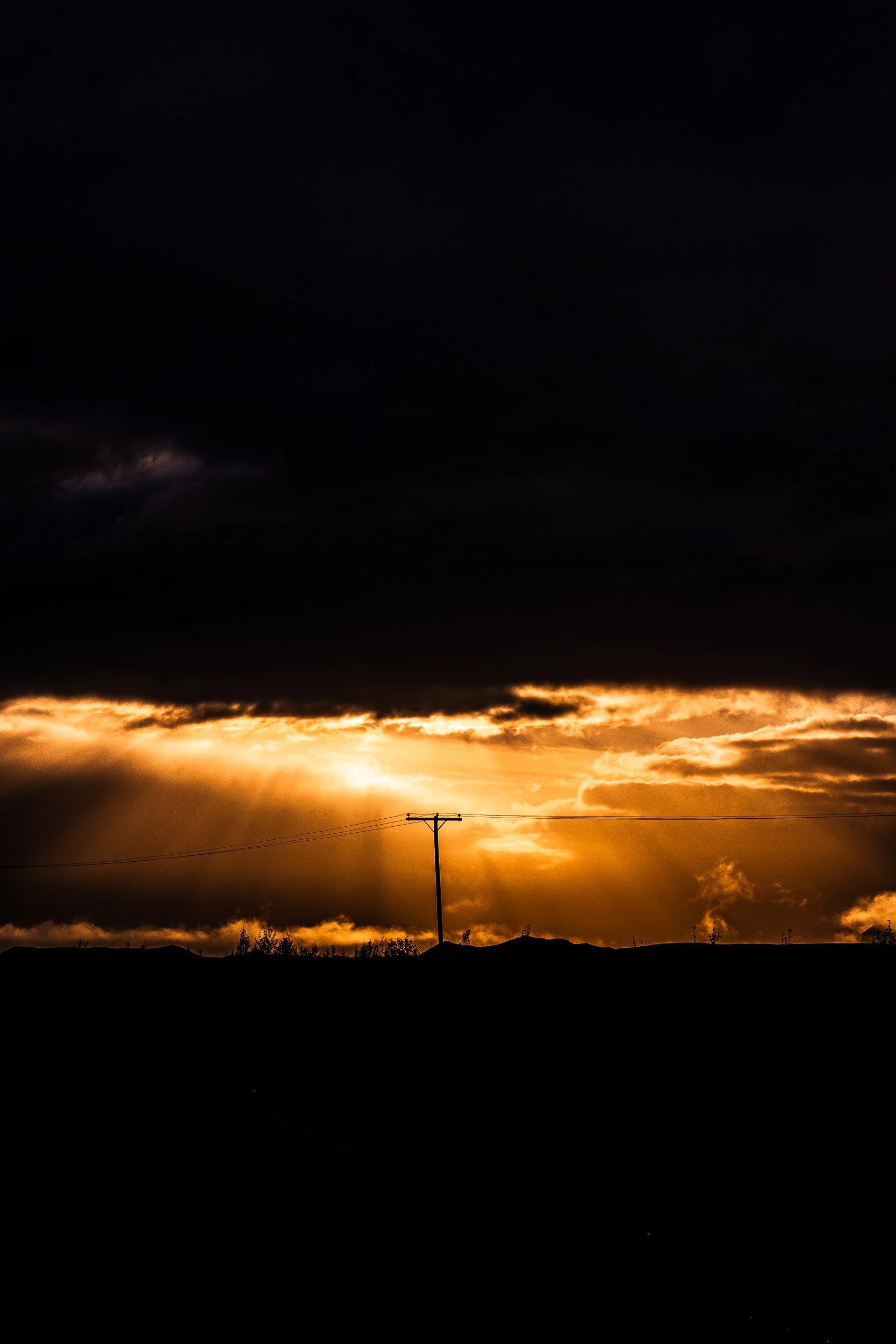 Dramatic golden sunset behind dark storm clouds, silhouetted landscape, and a utility pole with power lines