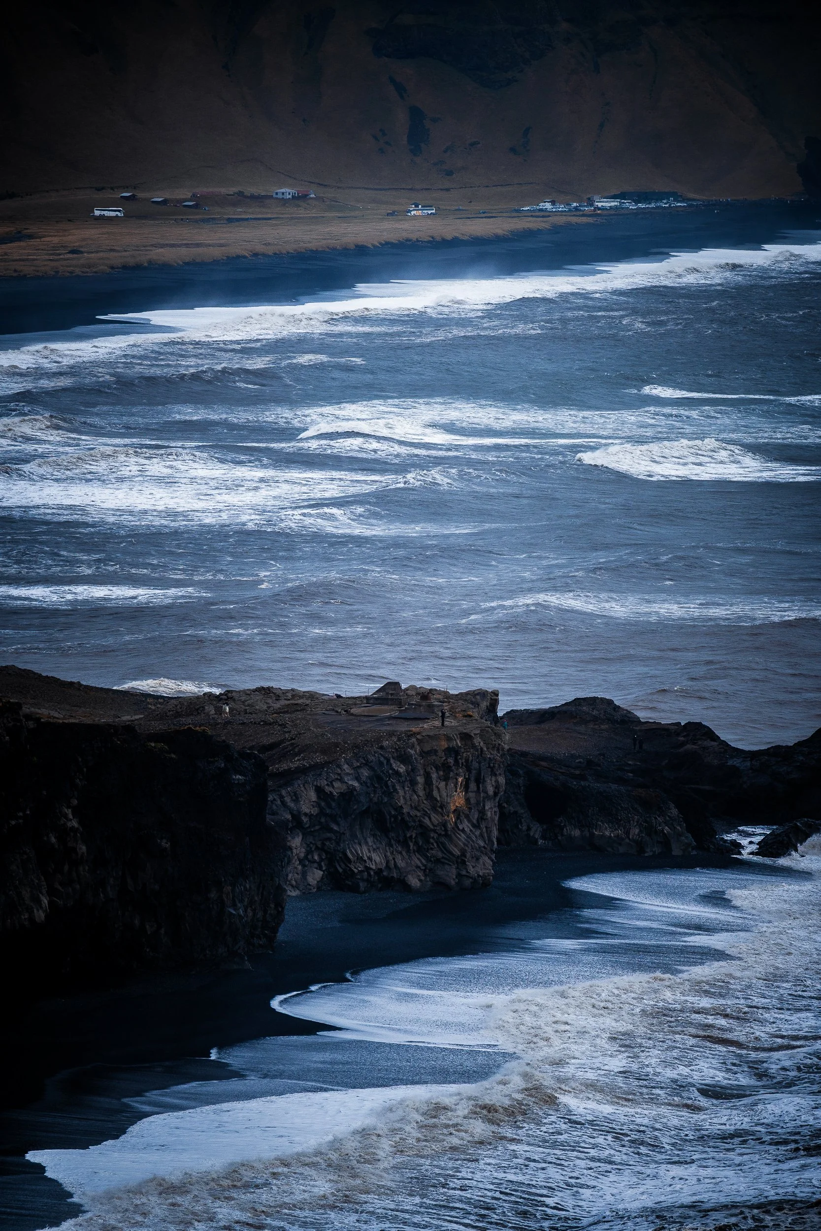 Stormy waves crashing onto a black-sand beach beneath rugged volcanic cliffs, with tour buses and a coastal settlement in the distance