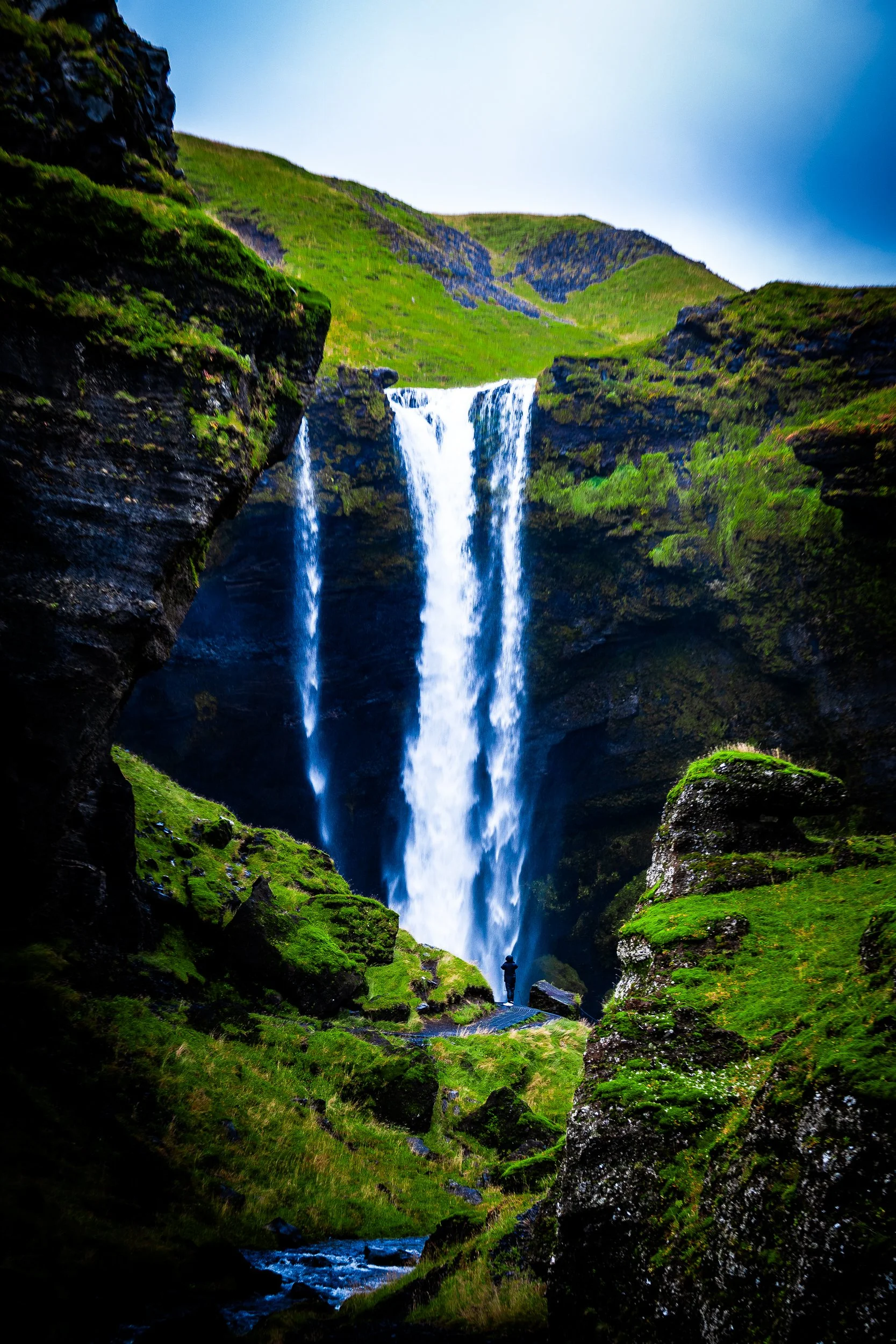 Tall waterfall cascading through moss-covered volcanic cliffs and green hills, with a small person viewing it below