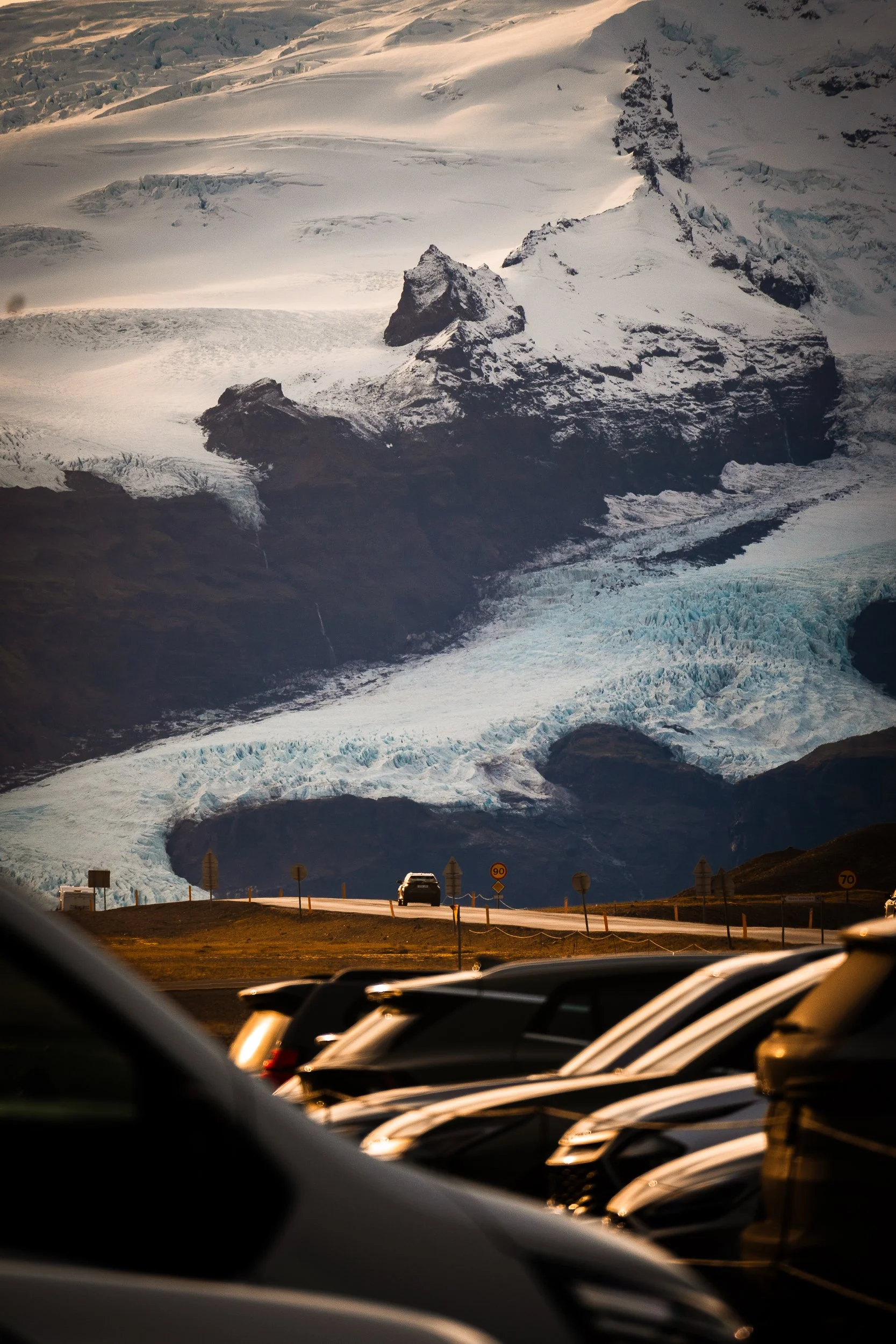 Aerial view of a snow-covered glacier and rugged volcanic peaks above a road with cars and speed limit signs.