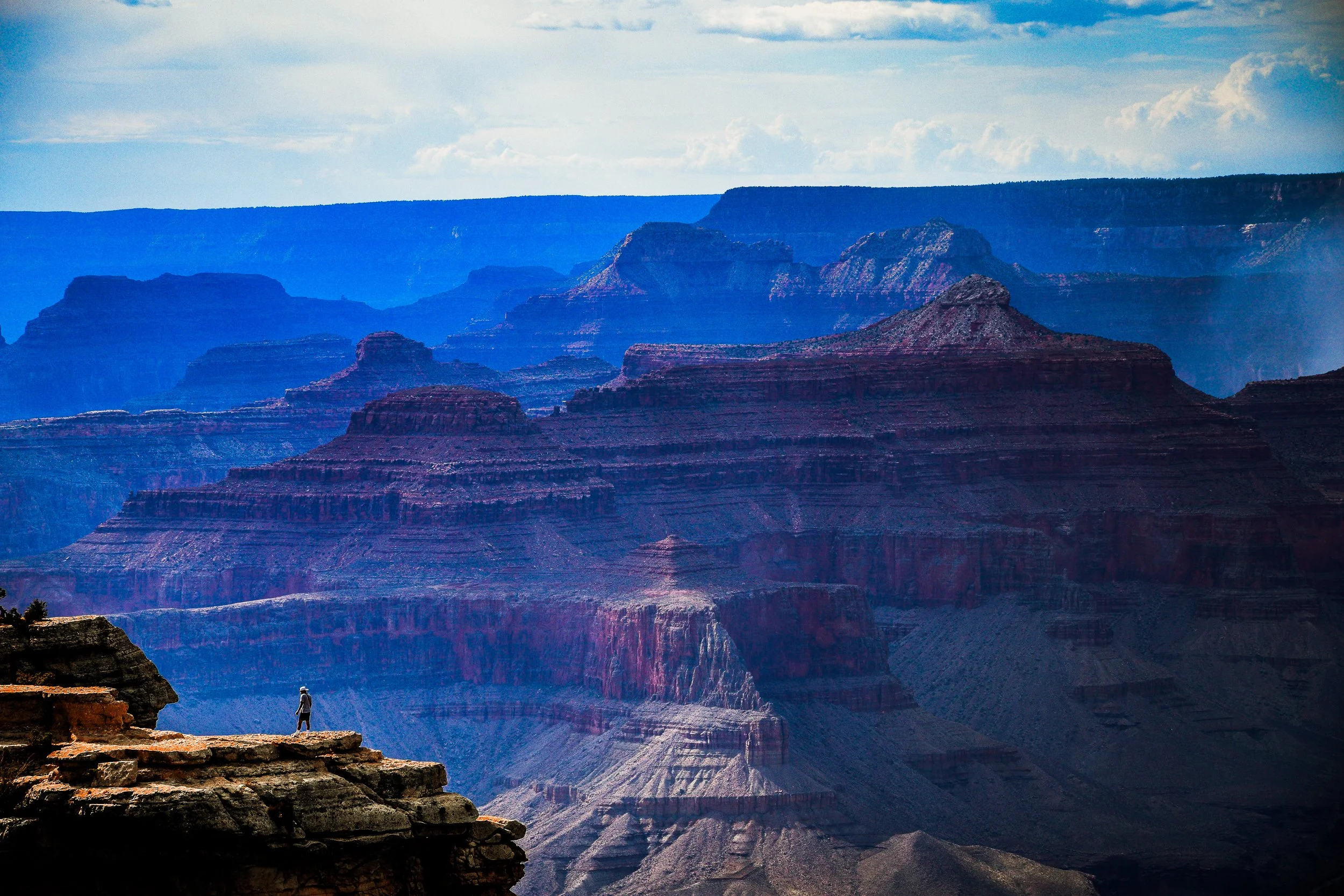 Layered red rock cliffs and mesas of the Grand Canyon beneath a partly cloudy sky, with a hiker standing on a foreground overlook