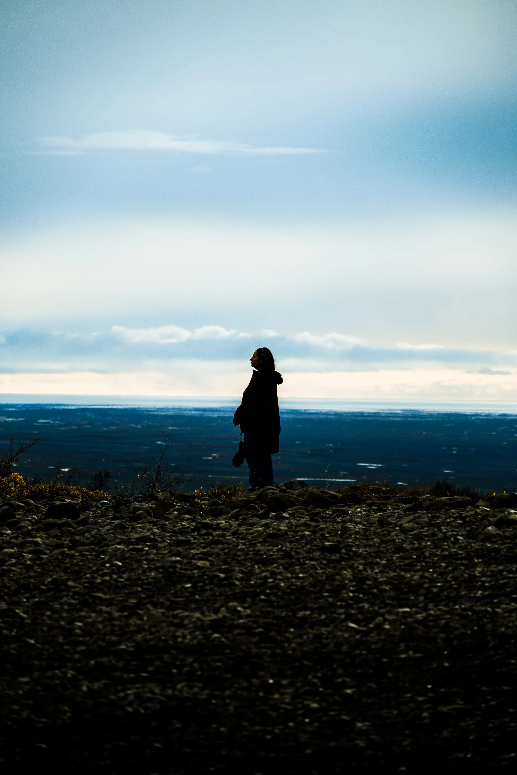 Silhouette of a person standing on a rocky shoreline overlooking the ocean beneath a cloudy blue sky