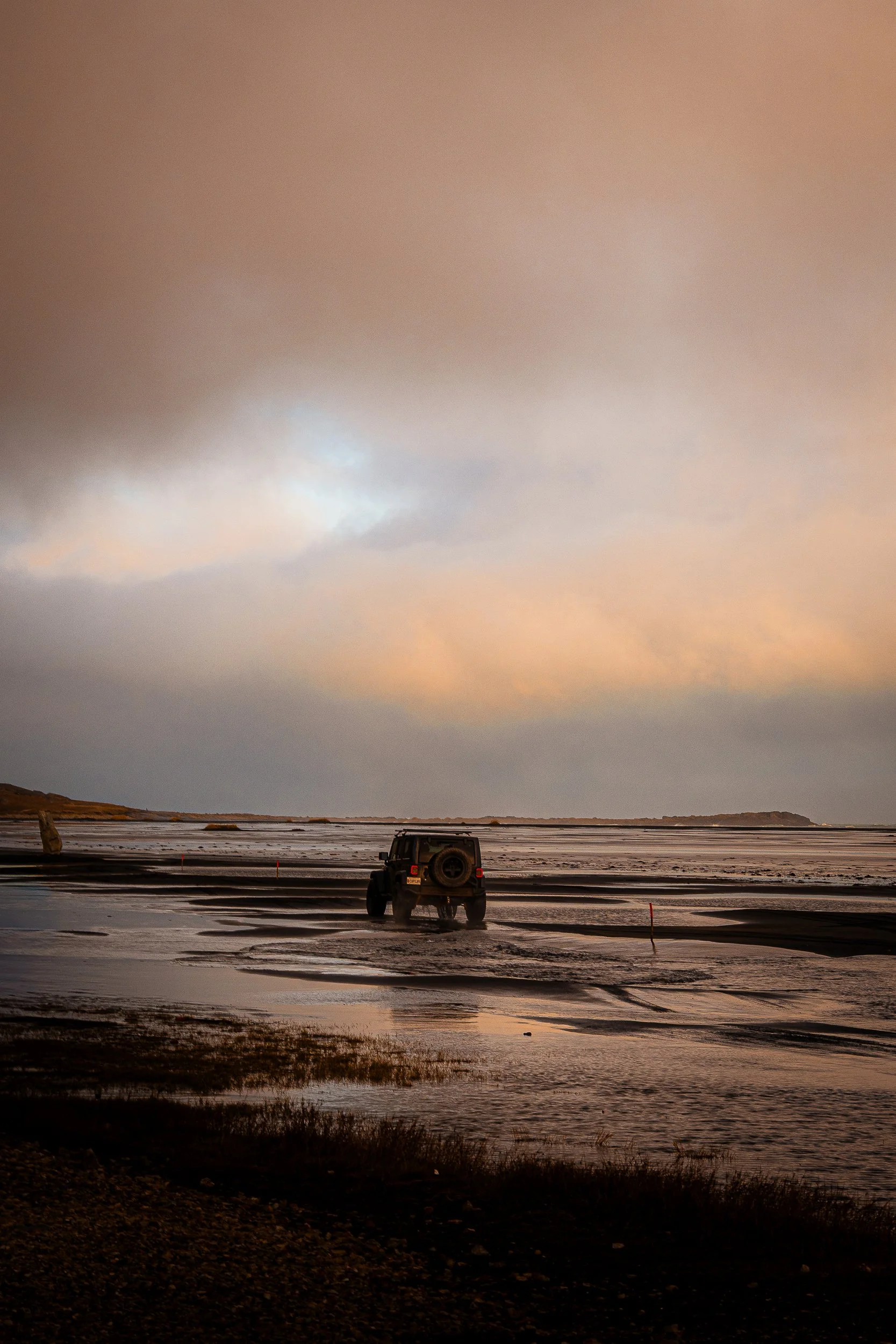 Off-road Jeep crossing a flat, muddy salt landscape beneath a dramatic sunset sky