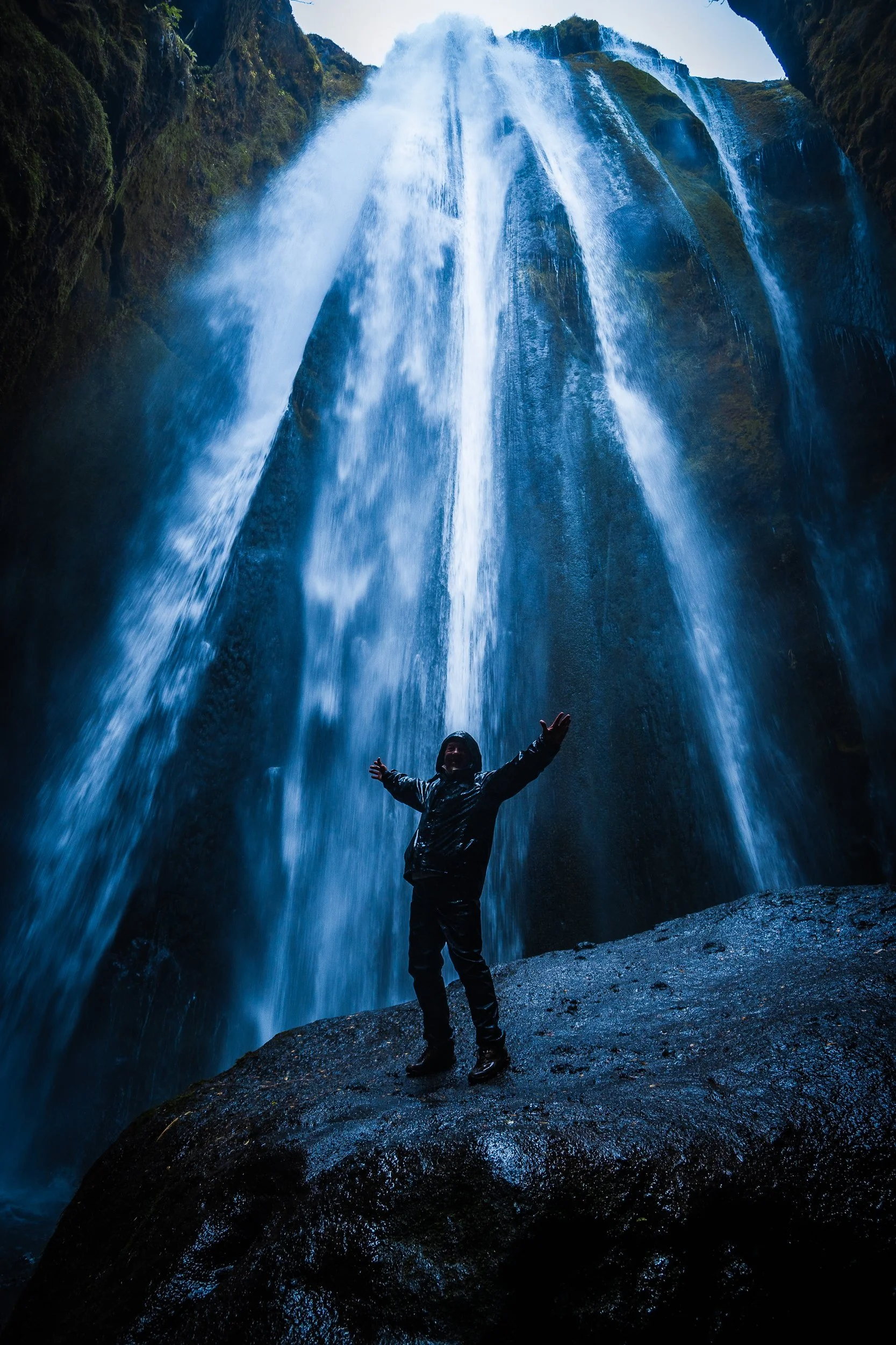 Traveler in a rain jacket standing beneath a massive cascading waterfall in a rocky canyon