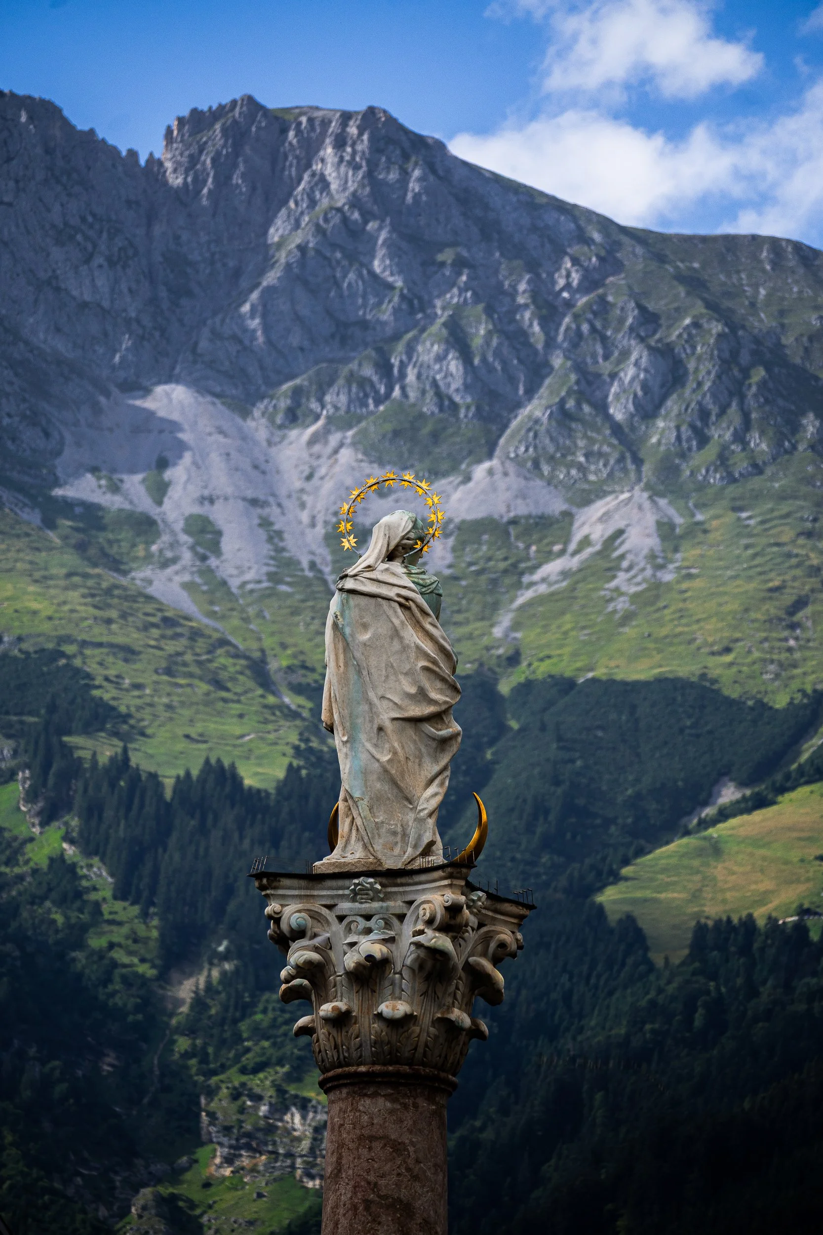 Statue of the Virgin Mary atop an ornate column with a golden halo, set against rugged green mountains and blue sky.