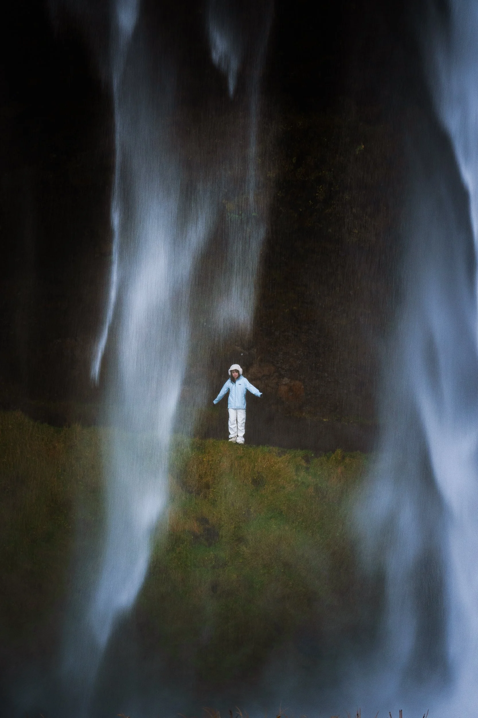 Person in white outdoor clothing standing beneath a tall waterfall in a forested landscape