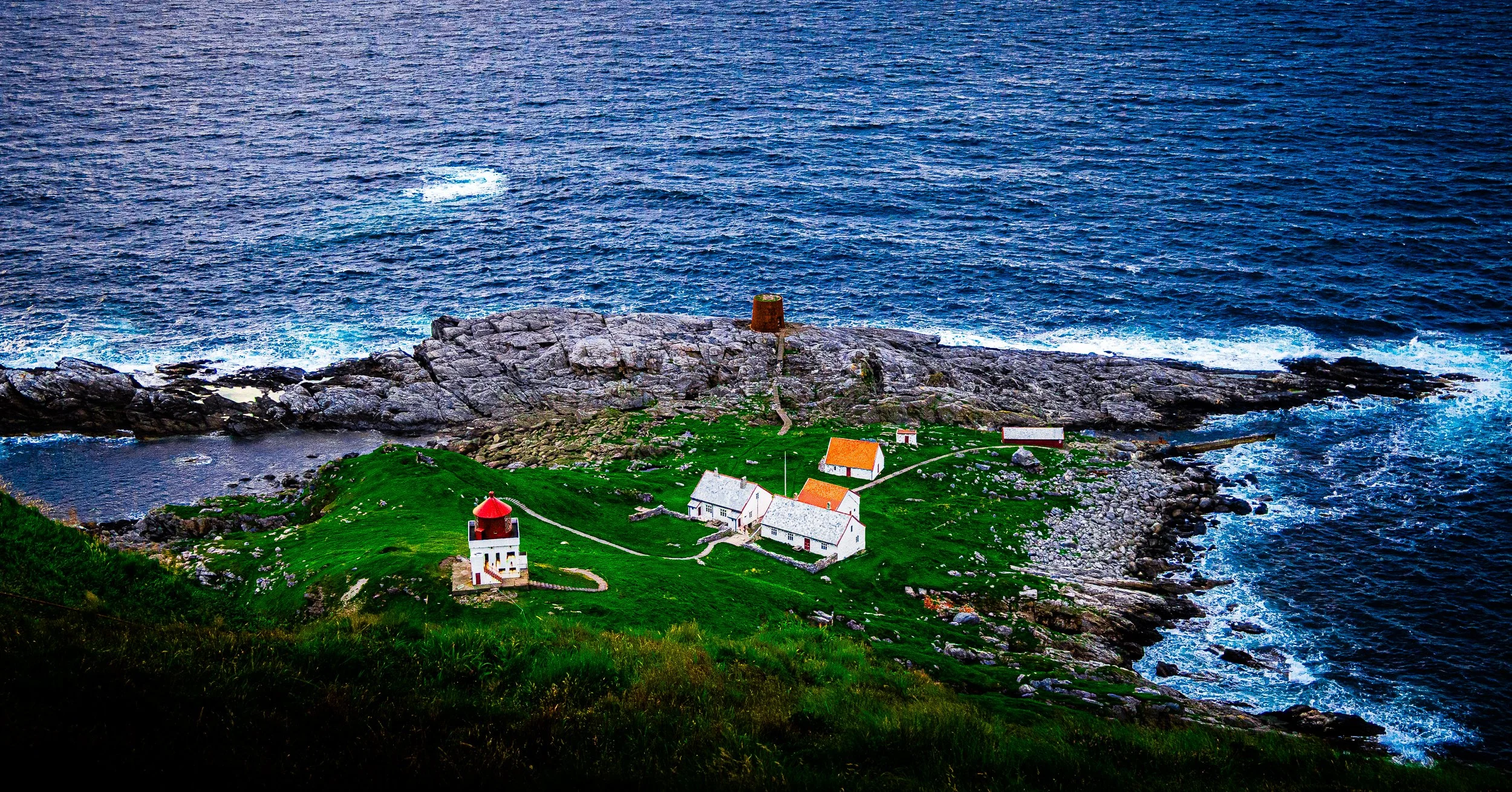Aerial view of a red-and-white lighthouse and buildings on a rocky green island surrounded by deep blue ocean waves.