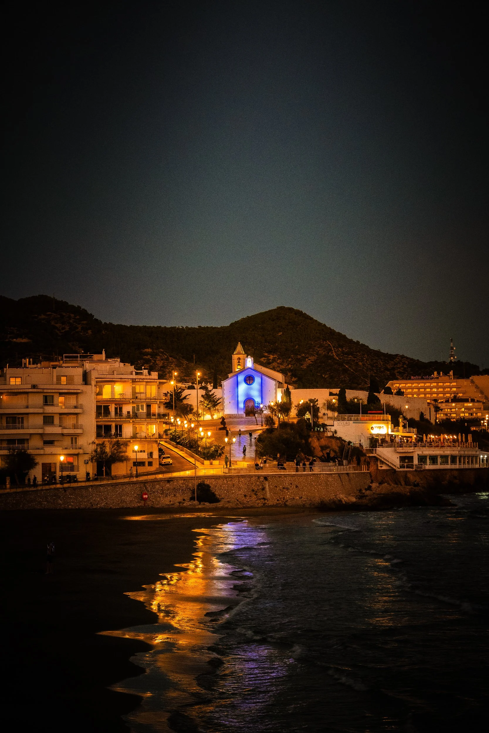 Nighttime coastal town with a blue-lit church, mountain backdrop, waterfront buildings, and streetlight reflections on the waves
