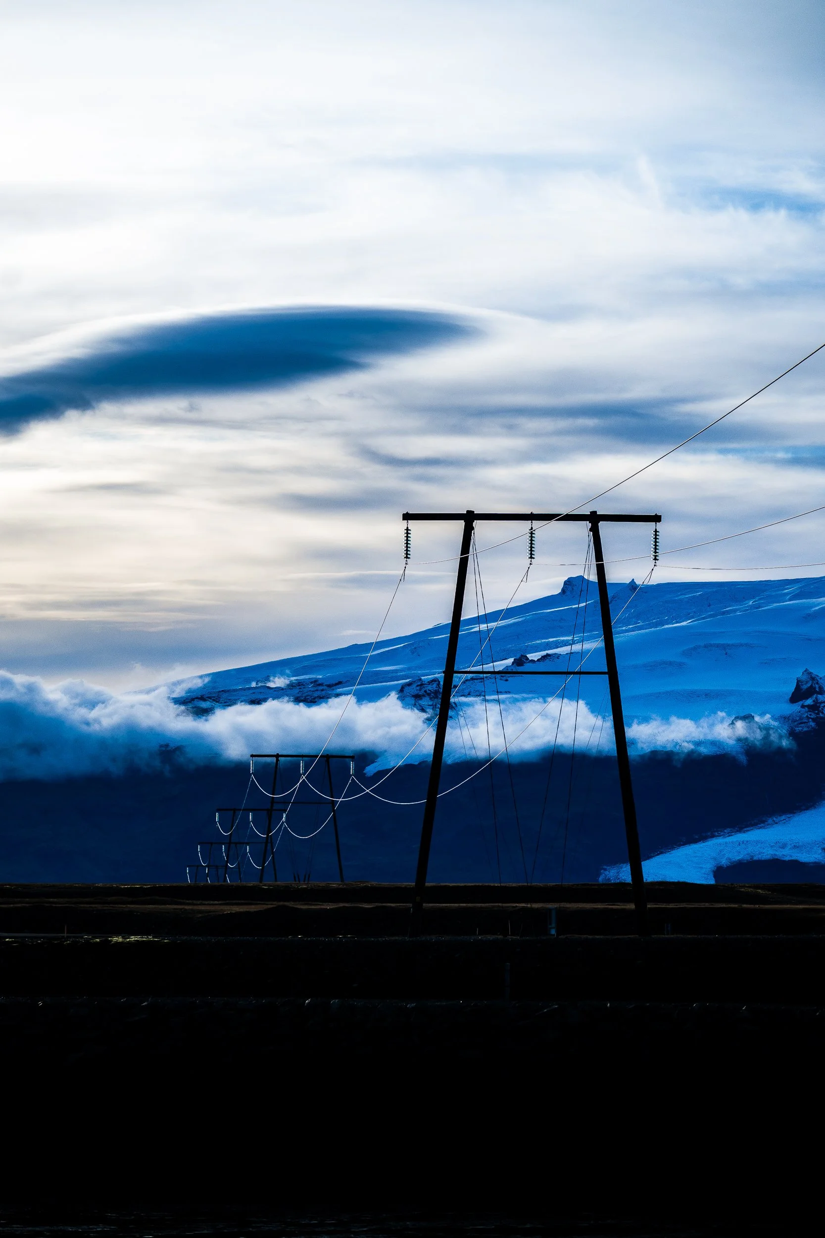 Snow-covered mountain landscape with power lines and utility poles beneath dramatic blue-and-white clouds