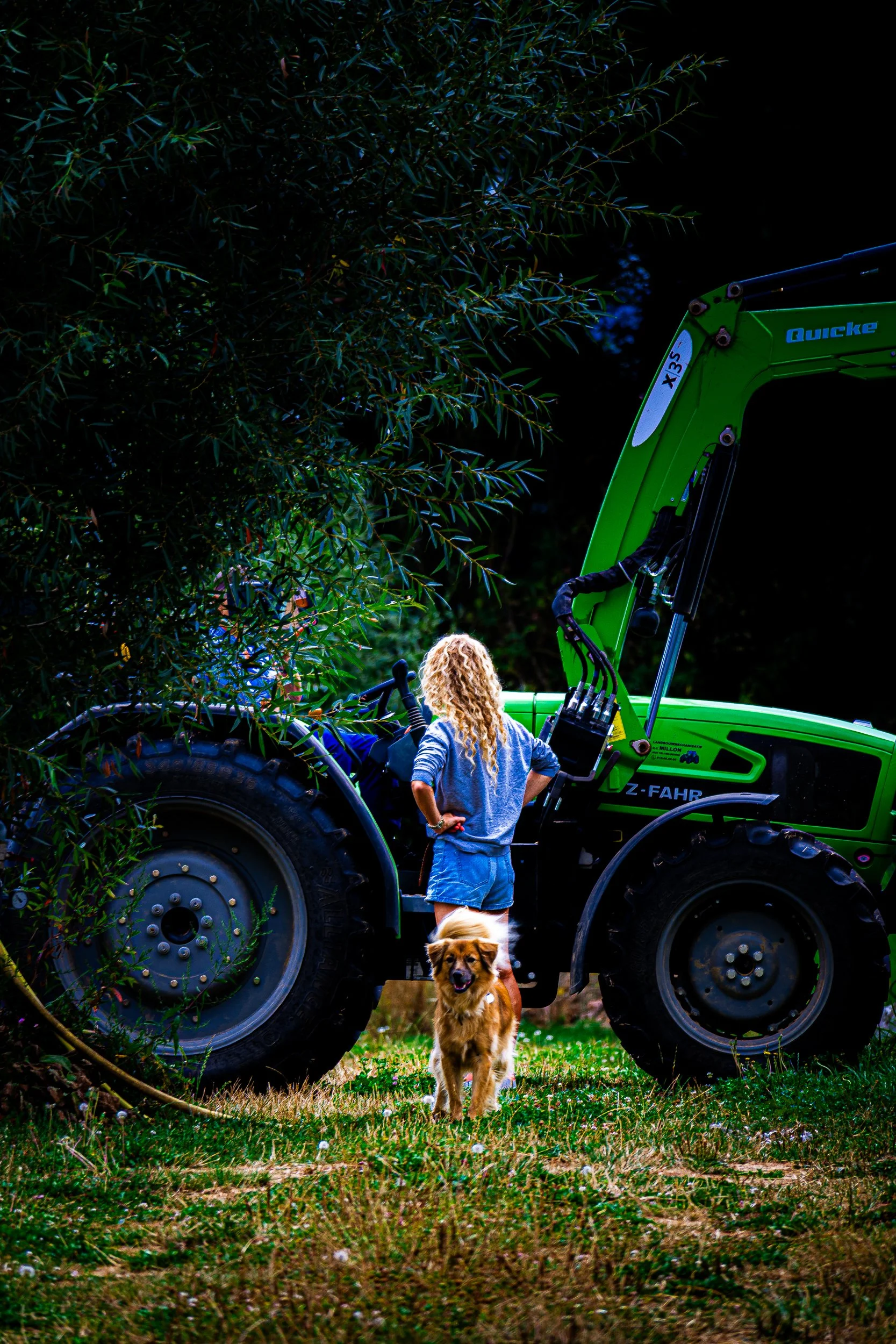 Woman standing beside a green Fendt tractor with a front loader while a dog sits in front, surrounded by leafy trees.