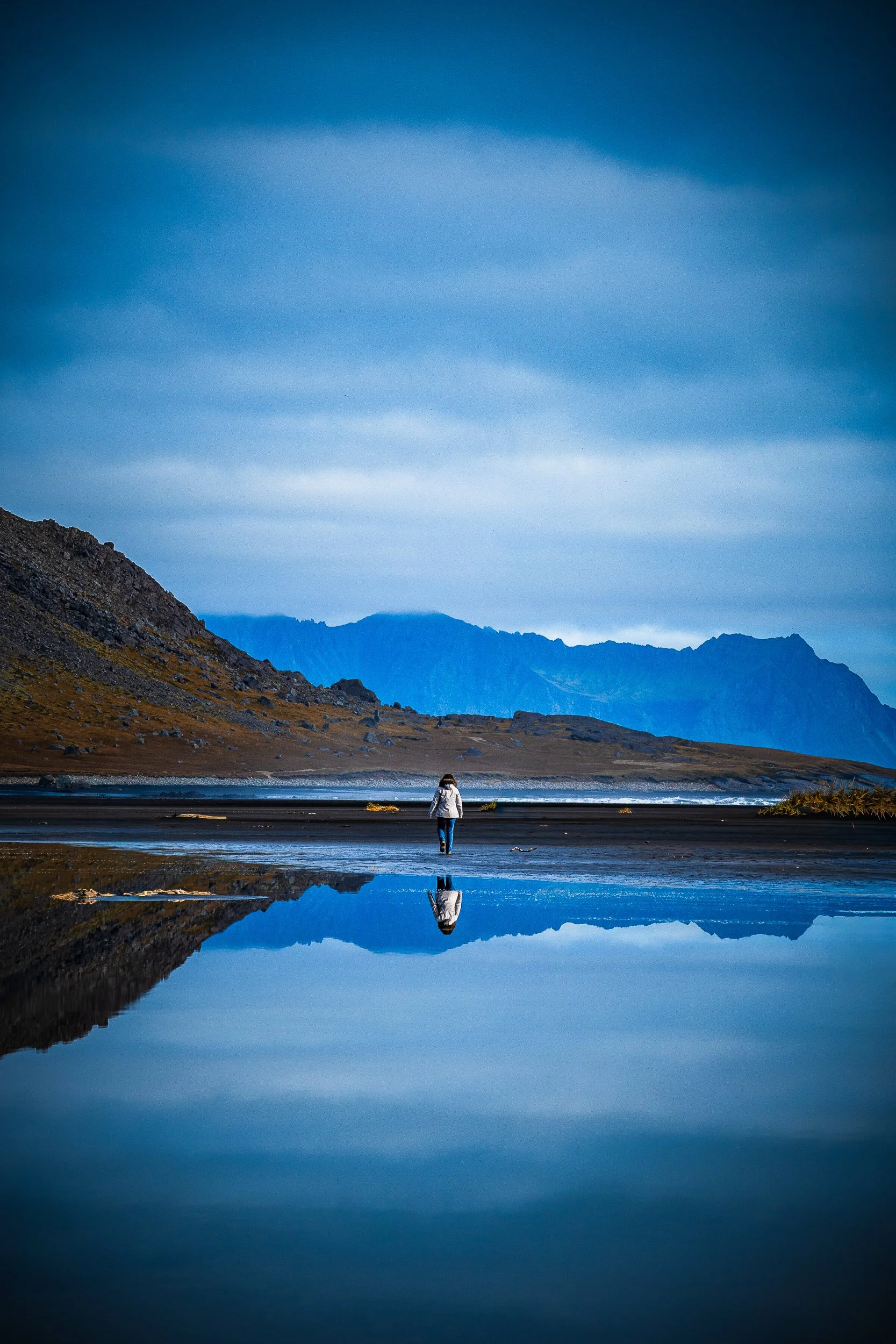 Person standing on a black sand beach beside a reflective pool, facing rugged mountains beneath a cloudy blue sky