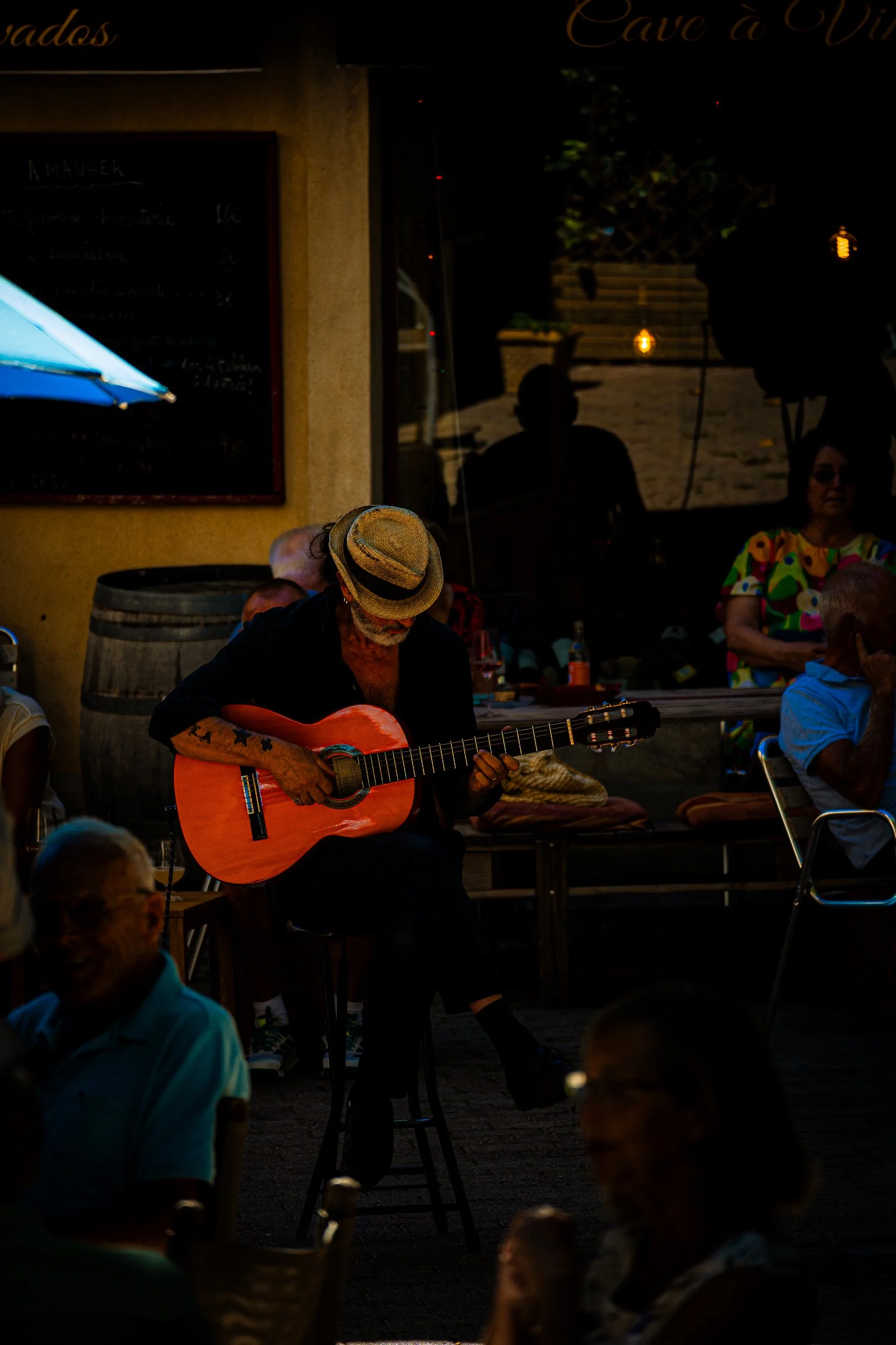 Street musician in a straw hat playing an acoustic guitar for an outdoor café crowd at night.