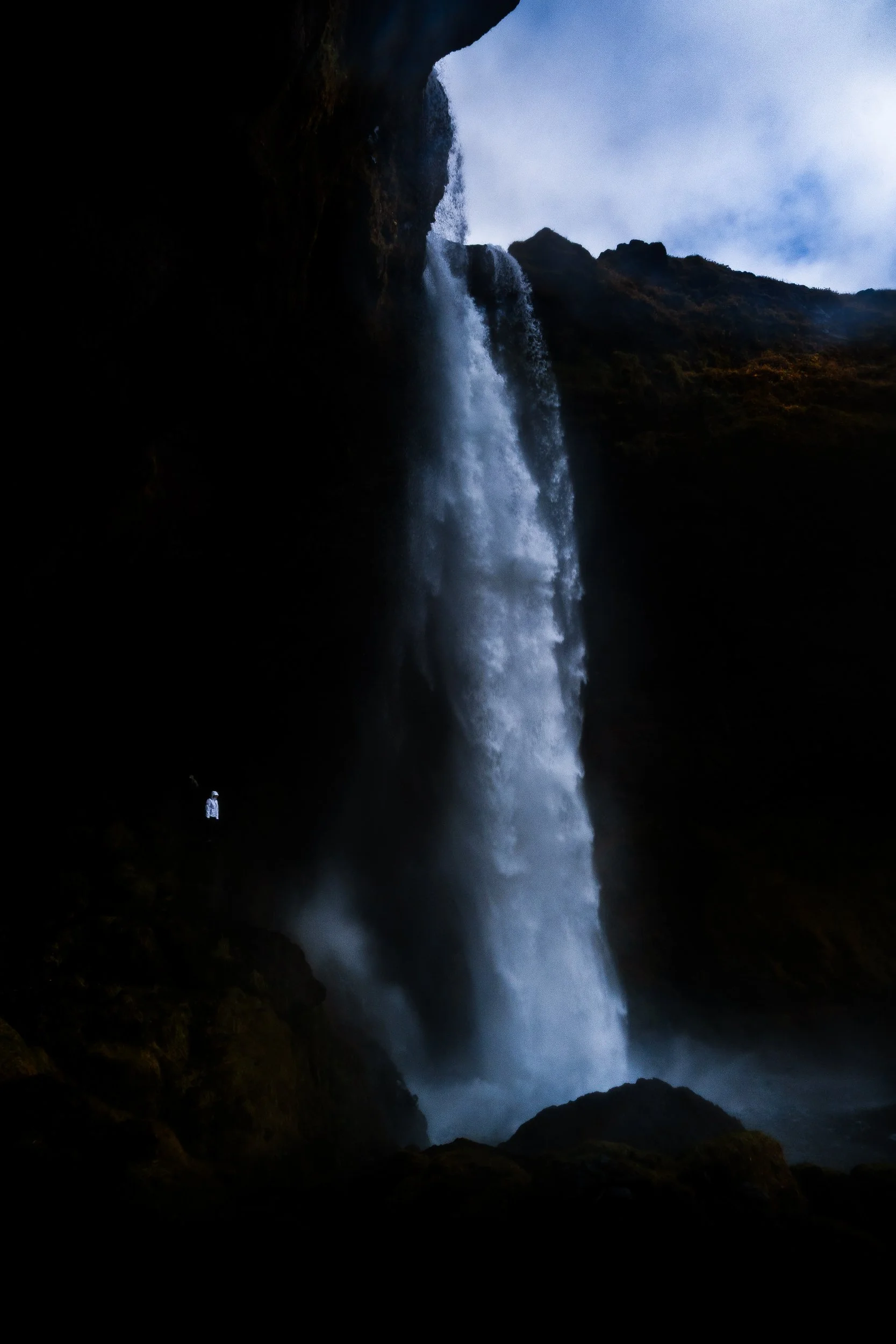 Tall waterfall plunging from a rocky cliff into a misty gorge, with a tiny hiker nearby