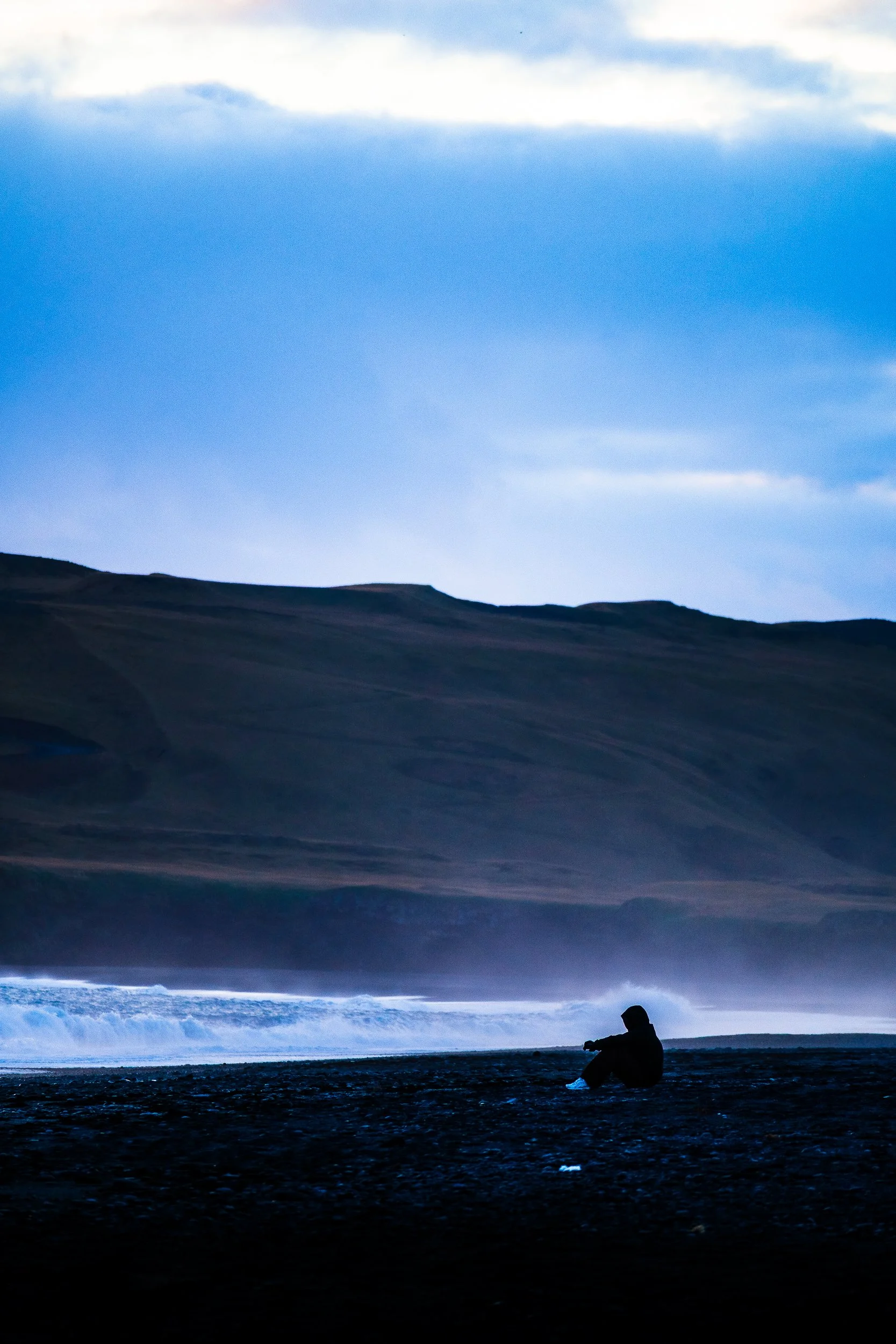 Silhouetted person sitting on a dark beach beside crashing waves, with barren hills and a cloudy blue sky in the background