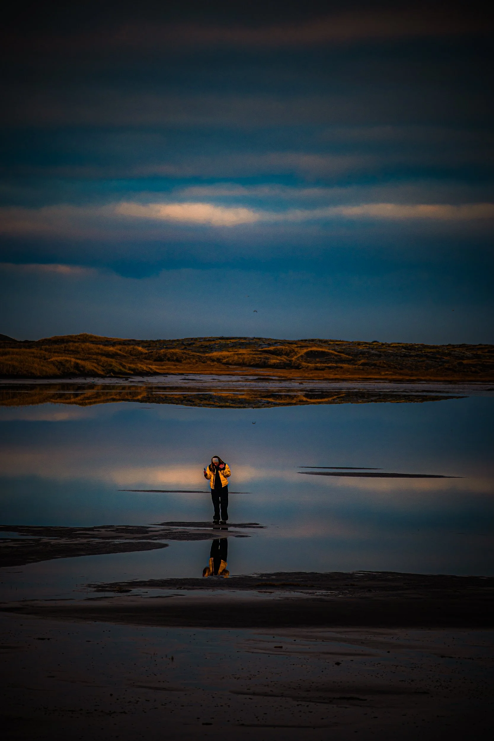 Person in a yellow jacket standing on a tidal flat, reflected in calm water beneath a dramatic cloudy sky