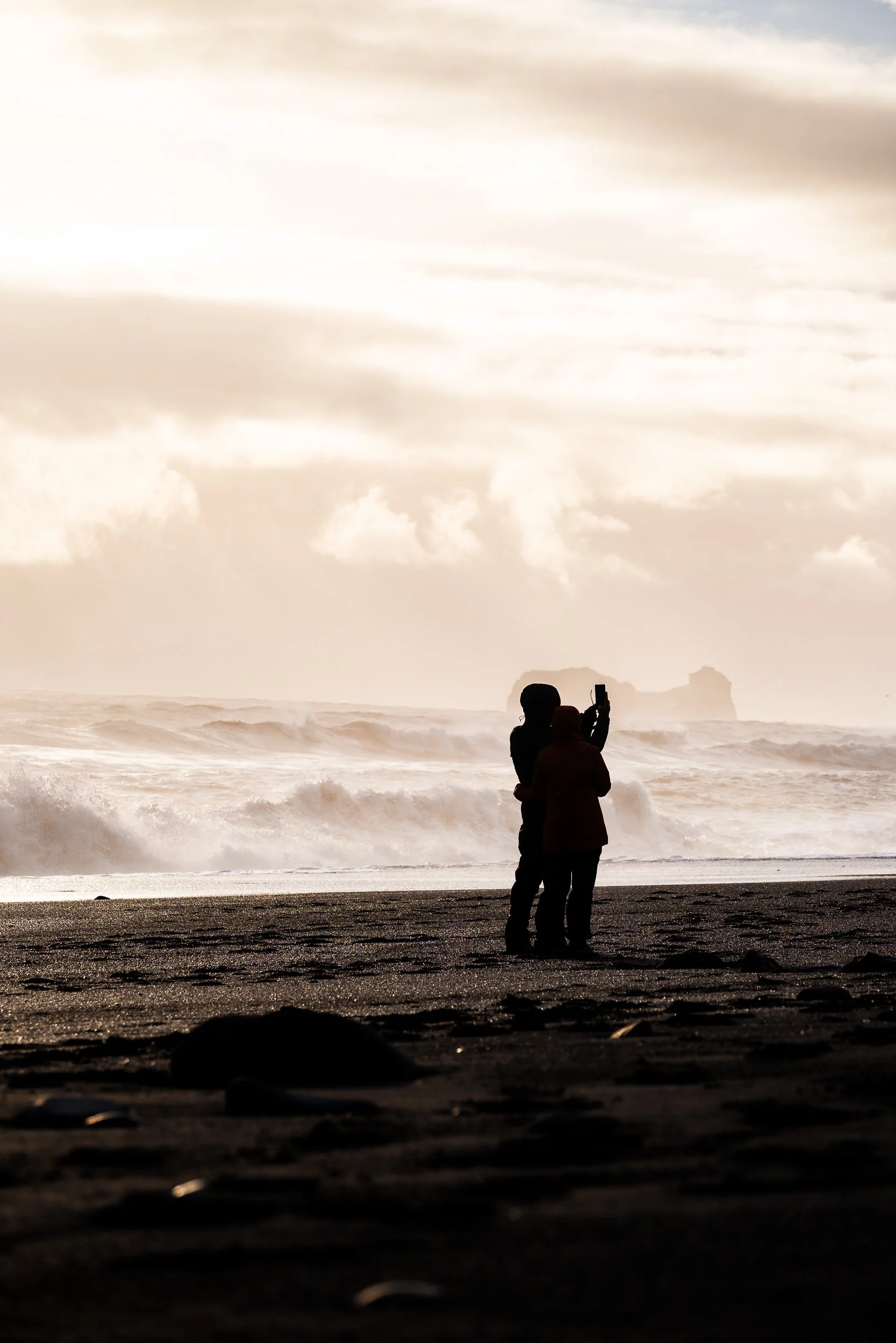 Two people photographing crashing waves from a black sand beach beneath a cloudy sky