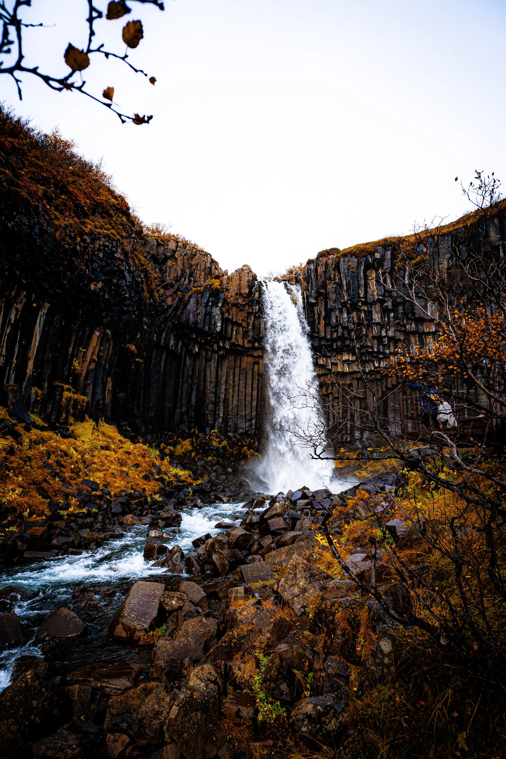 Waterfall cascading over basalt columns into a rocky stream surrounded by autumn foliage