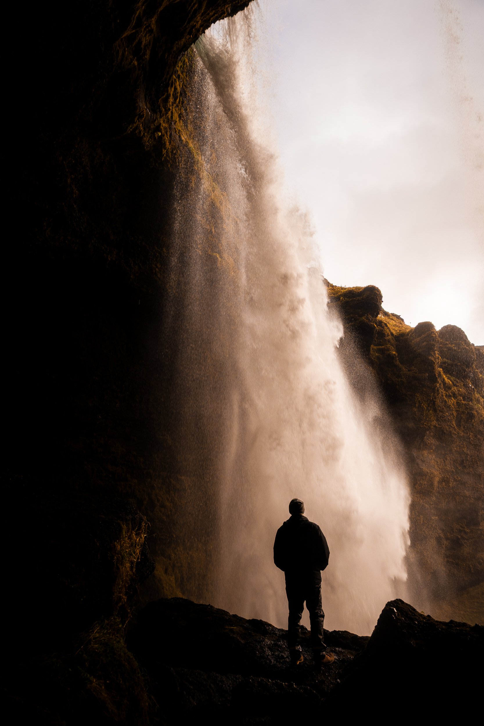 Person silhouetted beneath a powerful waterfall cascading through moss-covered cliffs