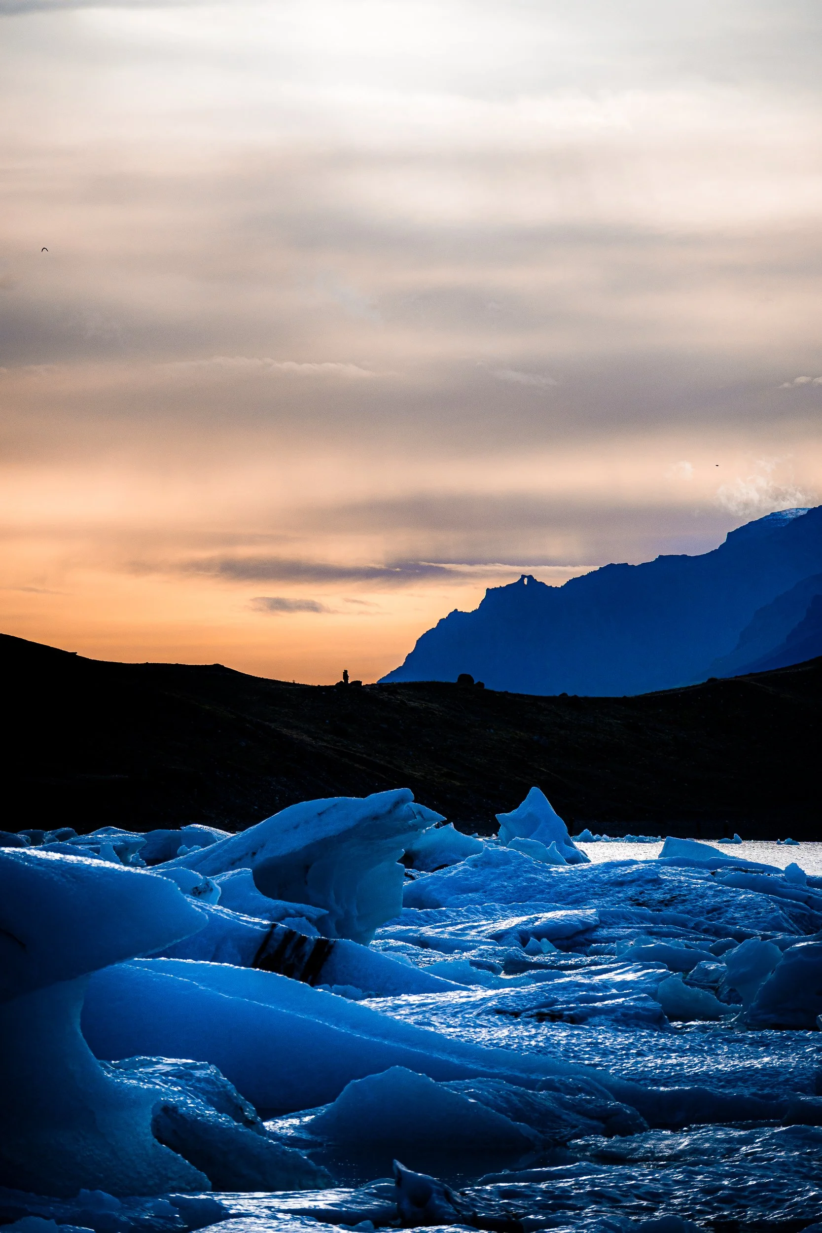 Blue ice formations floating in glacial water beneath a rugged mountain range at sunset