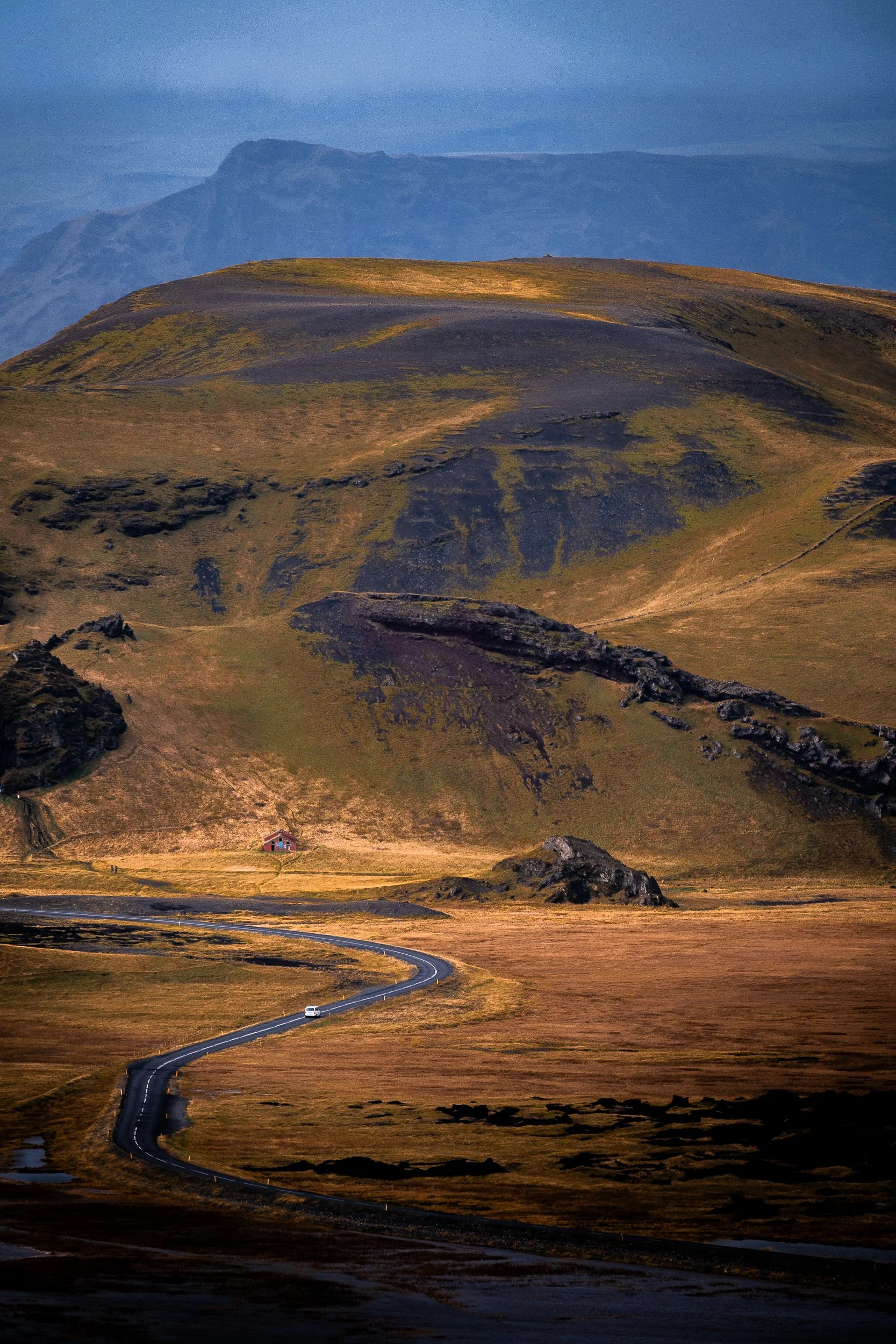 Winding Icelandic road through a volcanic mountain landscape with a small red cabin and distant blue mountains