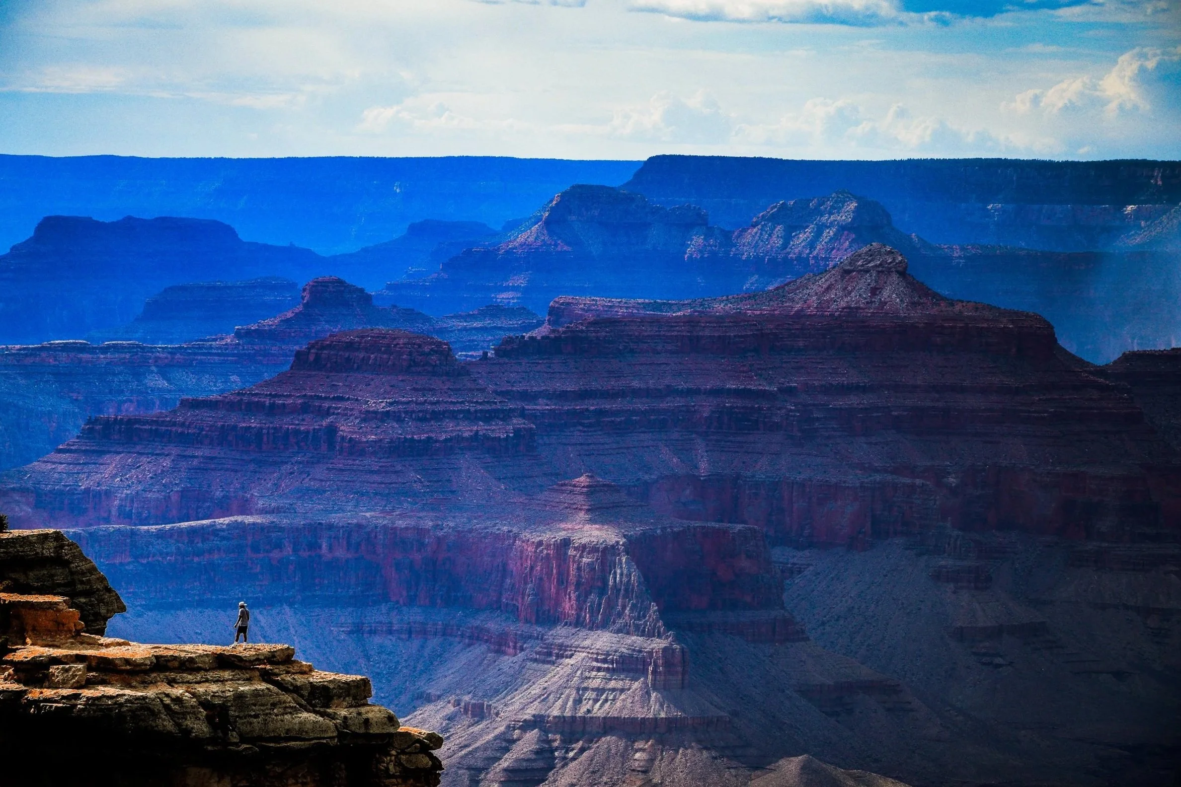 Layered red rock cliffs and mesas in the Grand Canyon under a partly cloudy sky, with a hiker on the foreground ledge