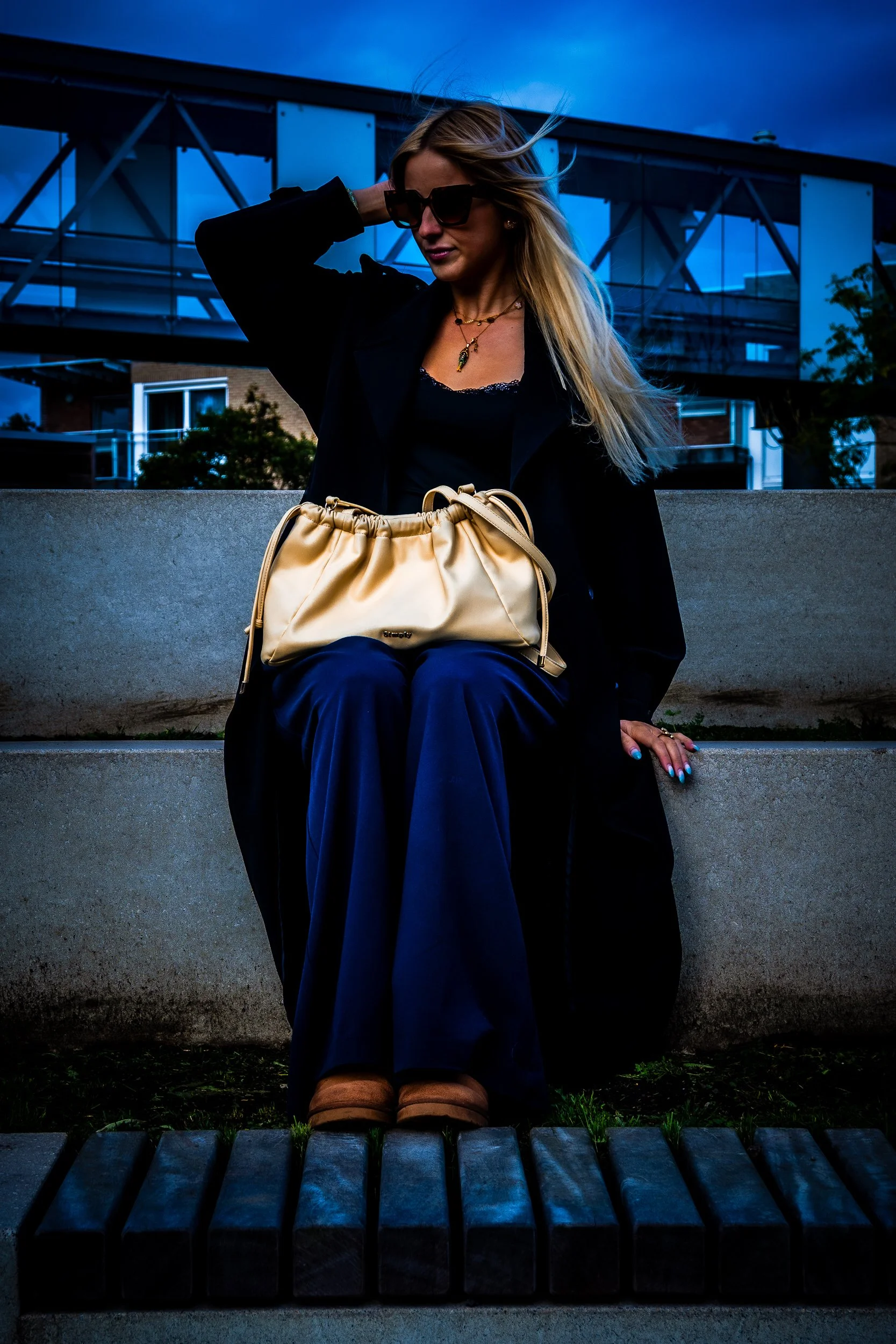 Woman wearing sunglasses and a black coat, sitting outdoors with a beige handbag