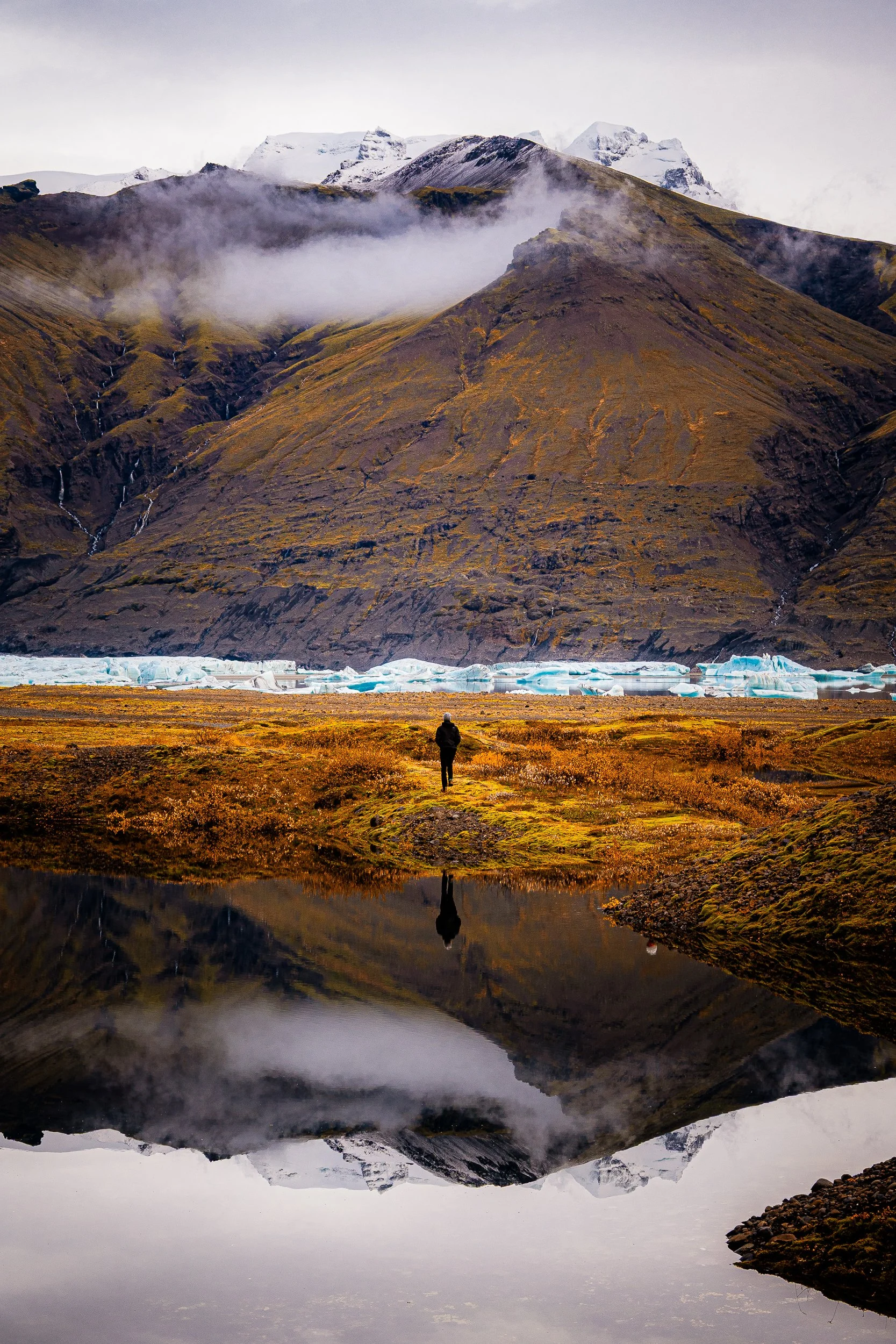 Lone hiker standing beside an iceberg-filled glacial lagoon beneath snow-covered mountains, reflected in calm water.