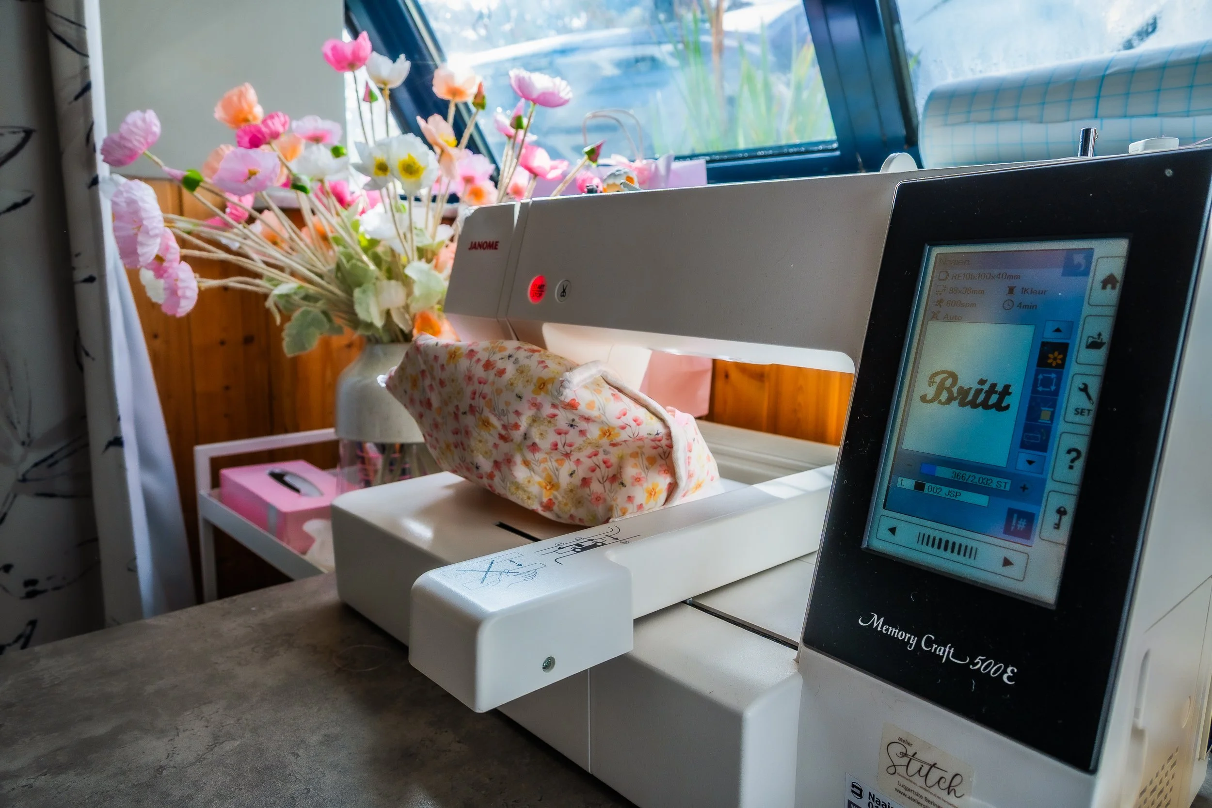 Janome Memory Craft sewing and embroidery machine stitching floral fabric beside a vase of colorful flowers.
