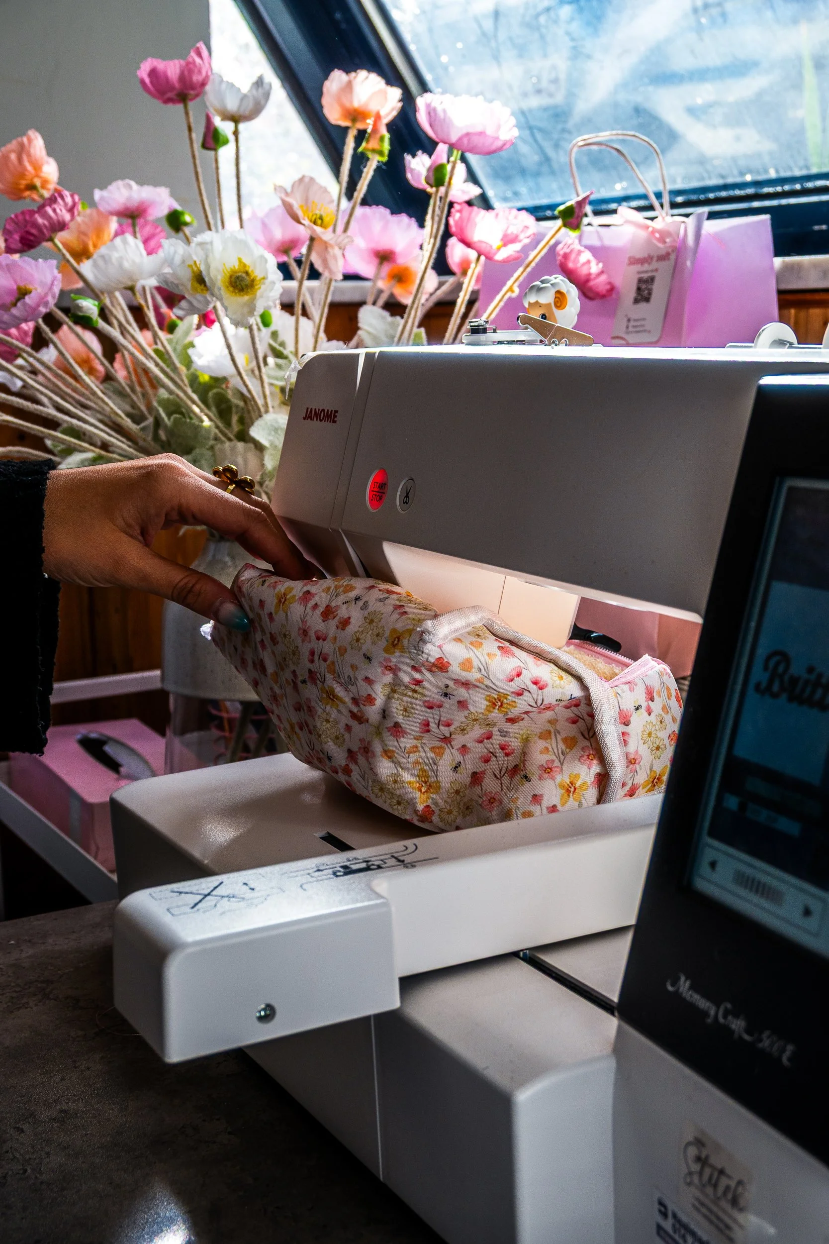 Person sewing floral fabric on a Janome sewing machine beside colorful flowers.