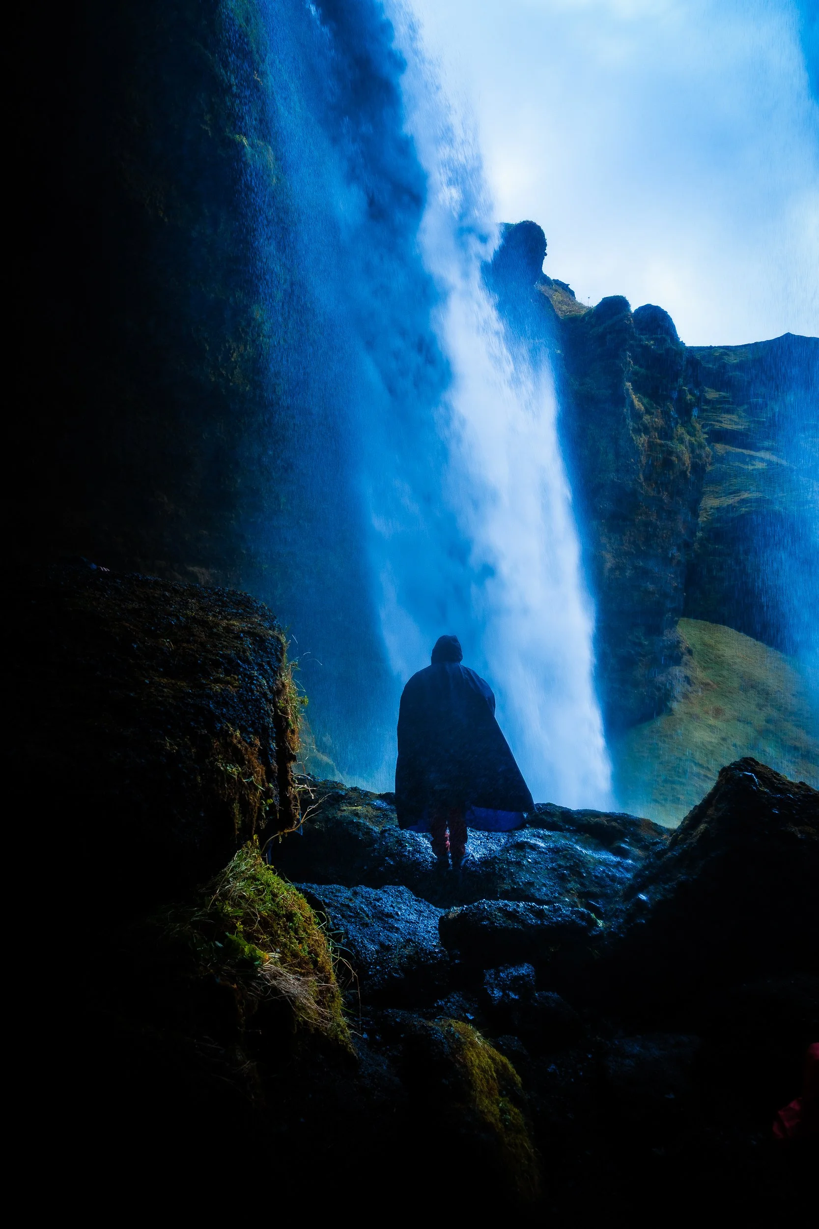 Person in a dark raincoat standing before a powerful waterfall in a mountainous landscape
