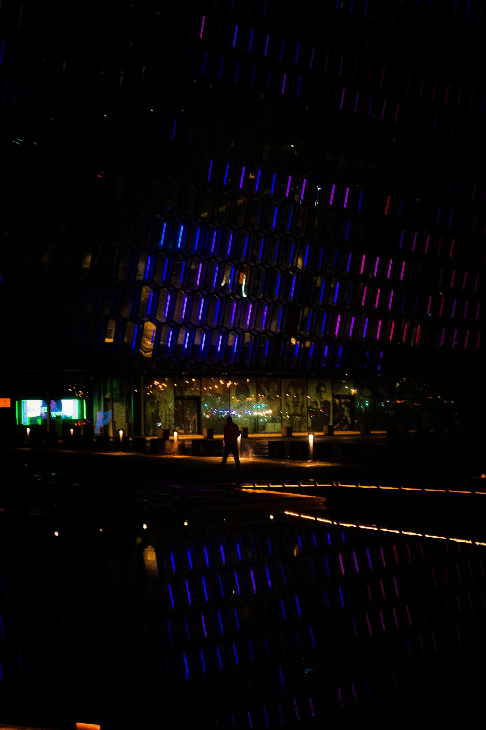 Nighttime view of a modern building covered in colorful blue and purple lights, reflected in a pool with a silhouetted person in front.