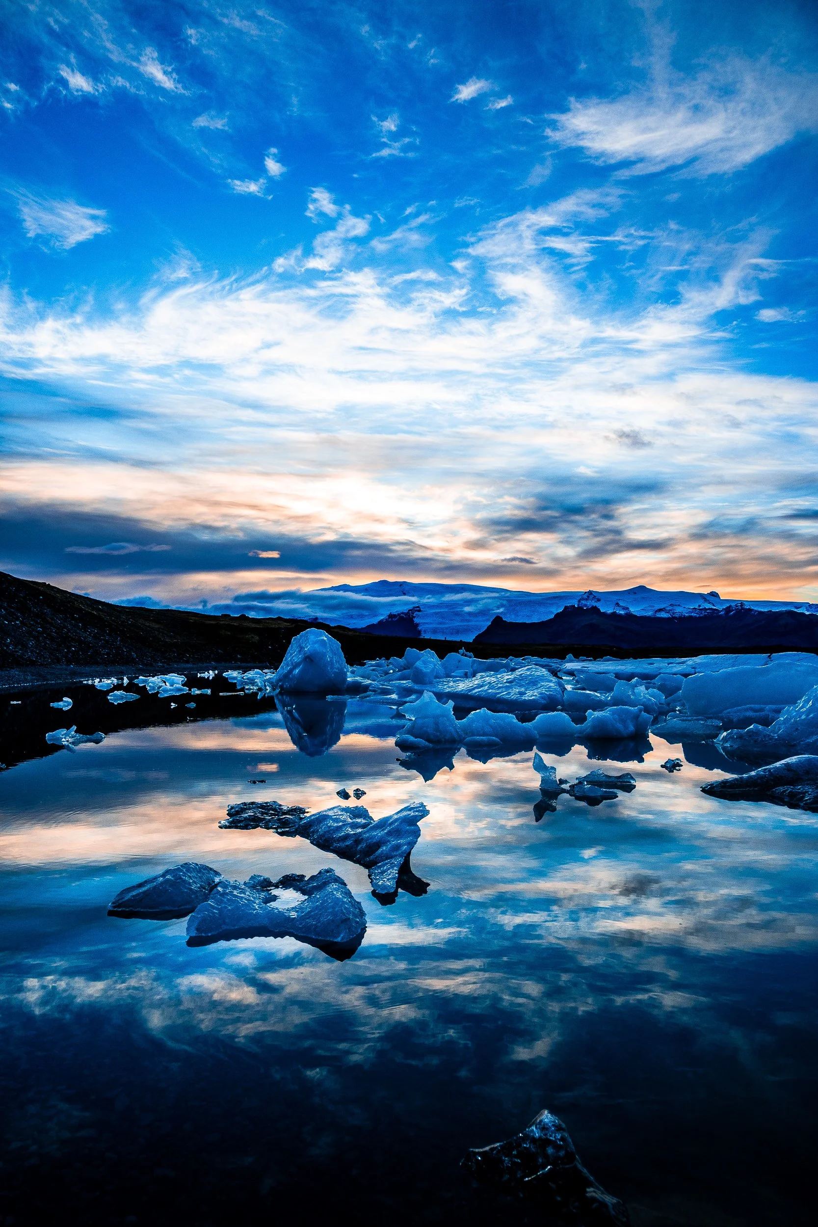 Blue icebergs floating in a glacial lagoon beneath a colorful sunset sky and snow-covered mountains