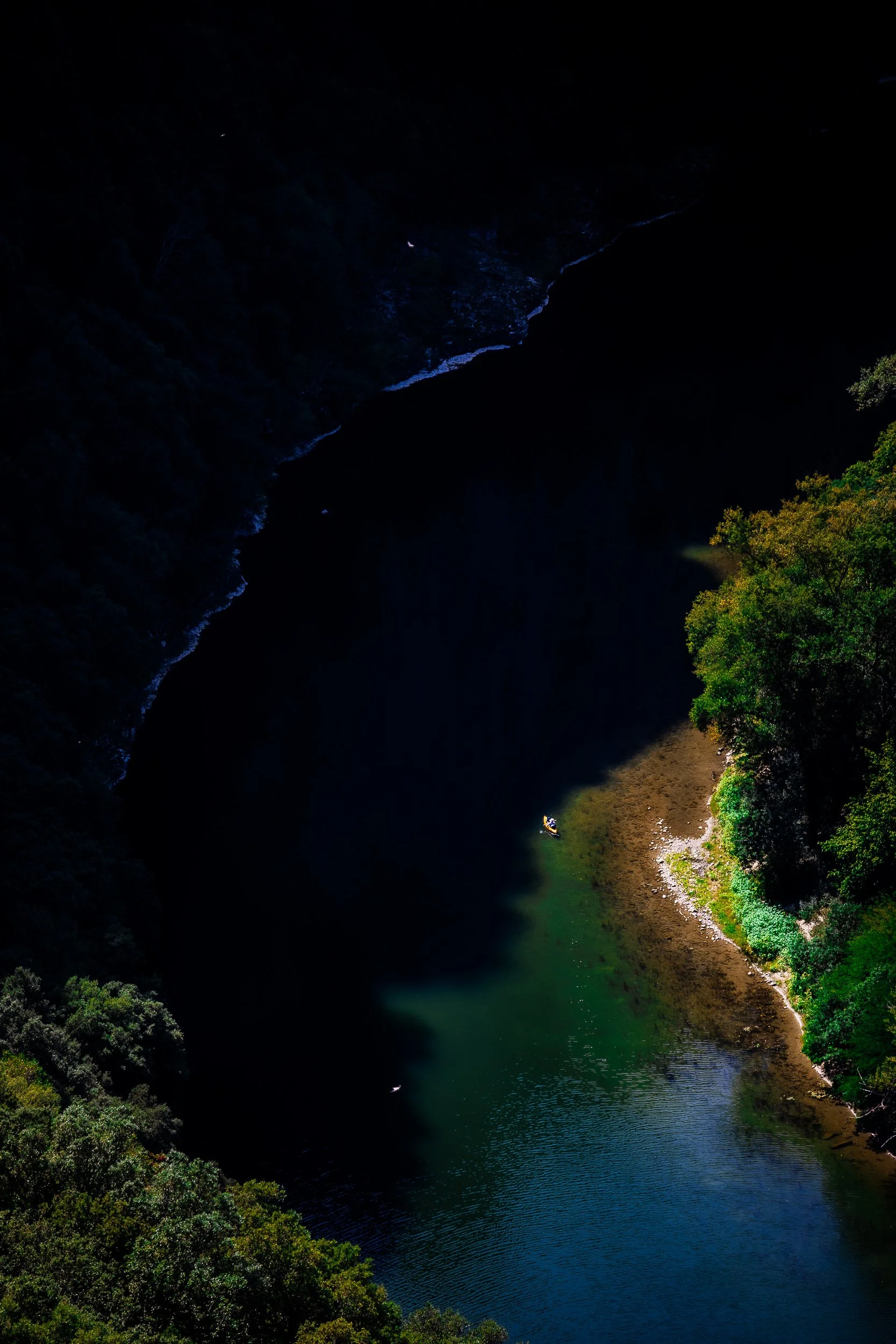 Aerial view of a forested river gorge with dark blue water, a rocky shoreline, and a small kayak near the bank