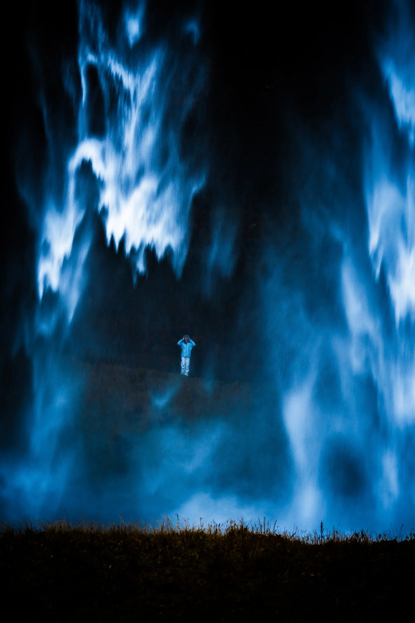 Person standing beneath a towering blue waterfall in a dark landscape