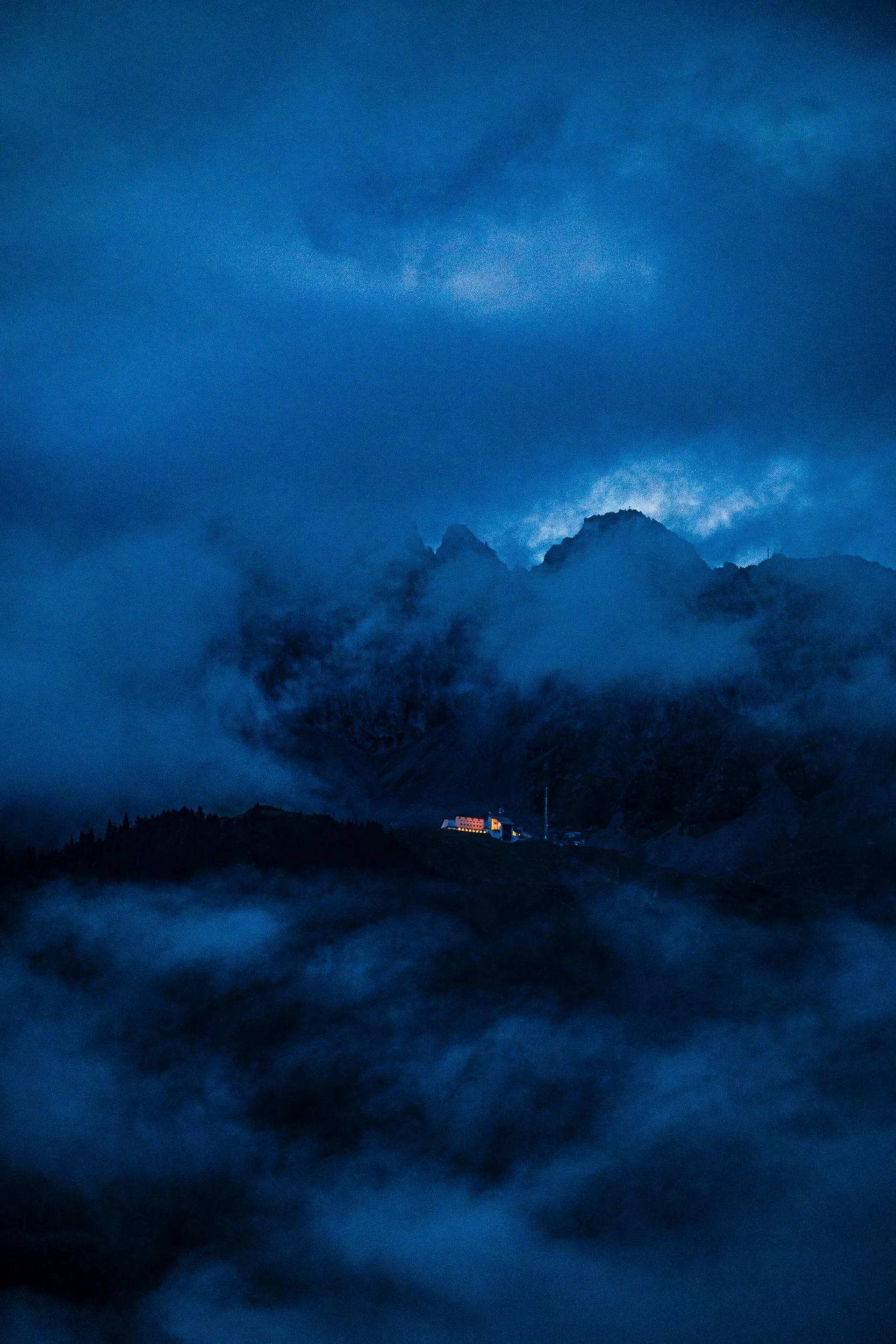 Dramatic blue twilight view of a cloud-covered mountain range with a warmly lit lodge in the foreground