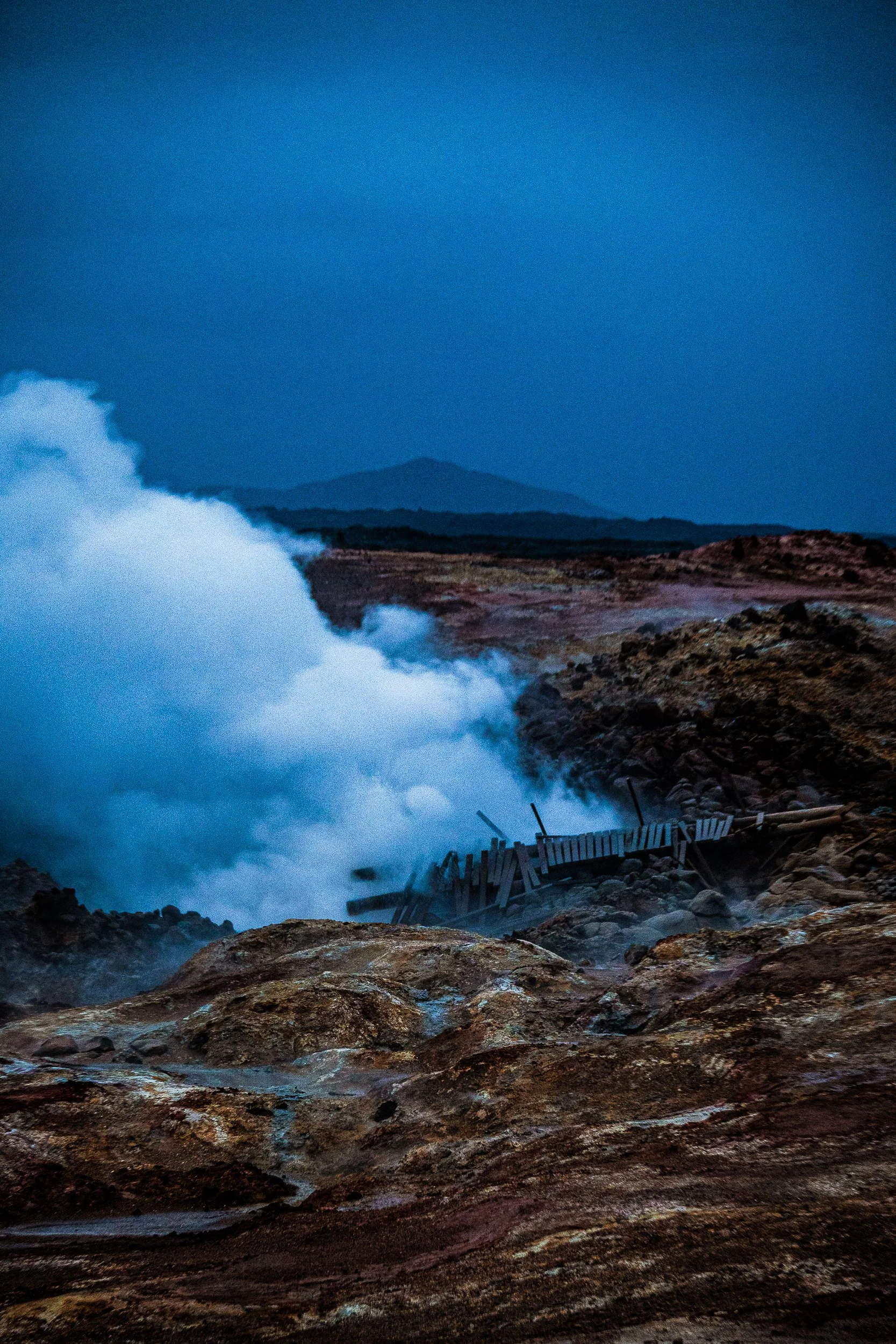 Large ocean wave crashing against a rocky volcanic coastline with a distant mountain under a blue sky