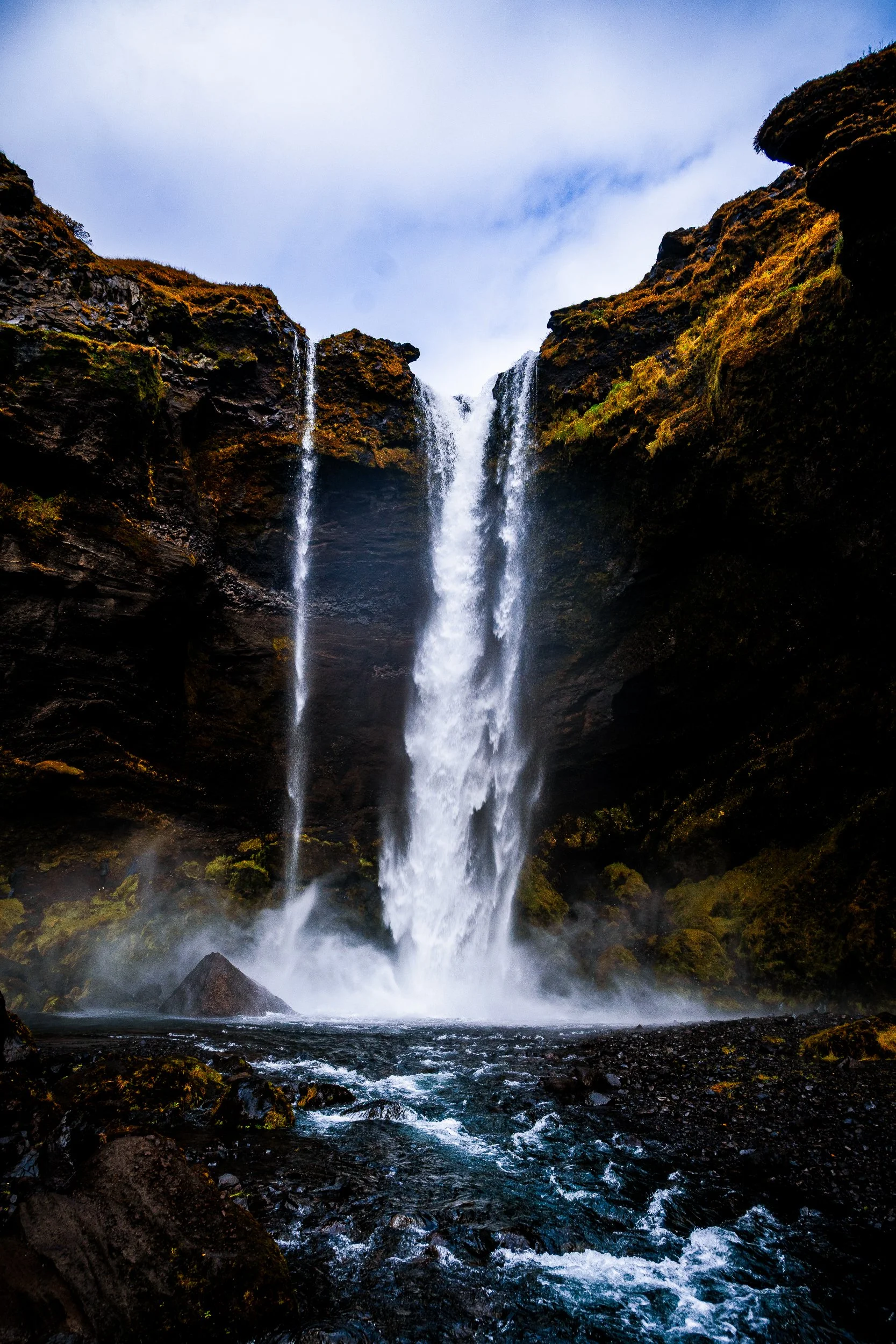 Tall waterfall cascading between moss-covered dark cliffs into a rocky stream under a bright blue sky