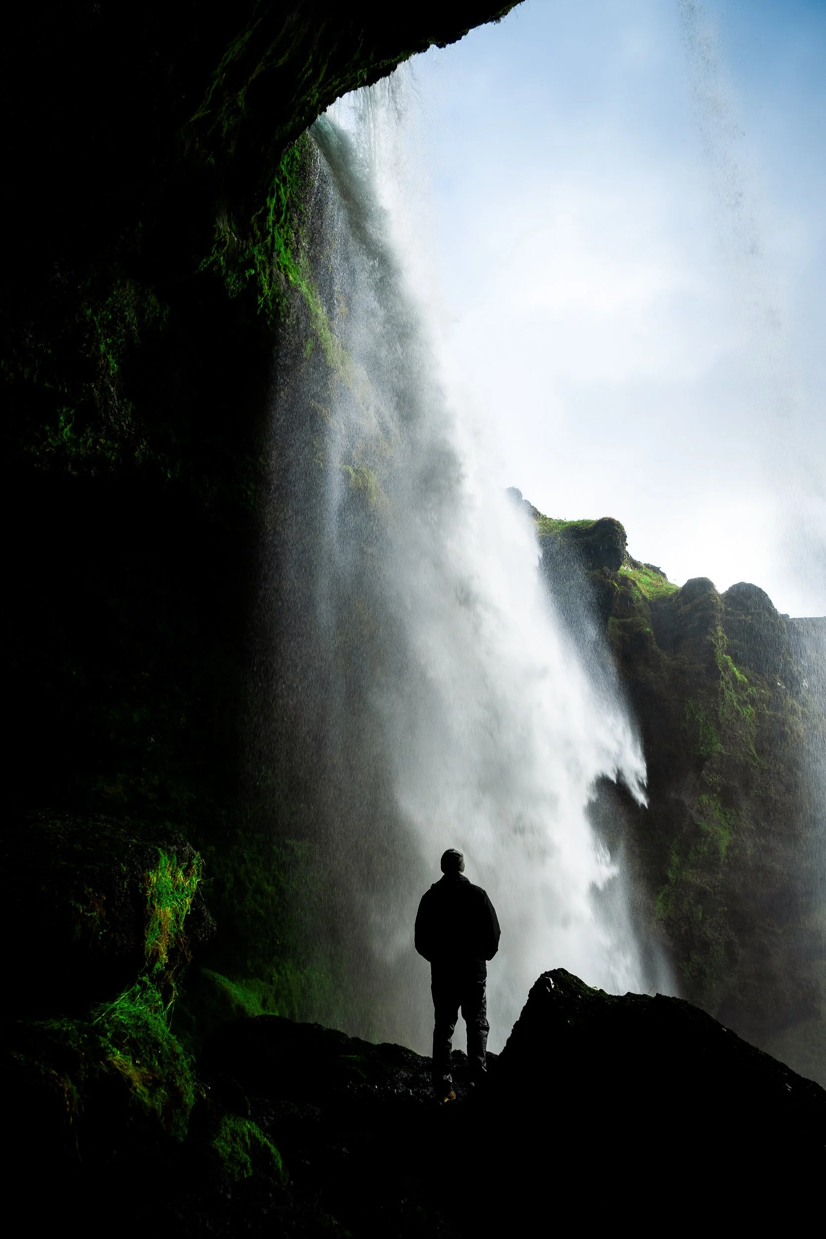 Person standing beneath a powerful waterfall inside a moss-covered cave